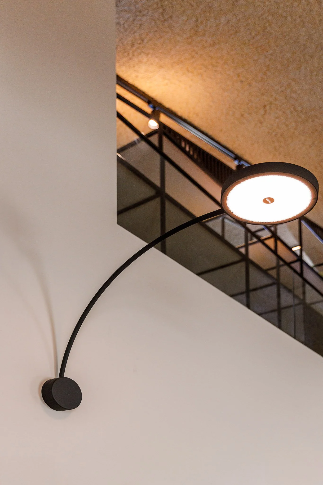 Luminária de parede moderna preta com braço curvado, fixada na parede branca, iluminando o ambiente com luz quente, no interior de um edifício com teto de madeira e varanda com grades pretas no fundo.