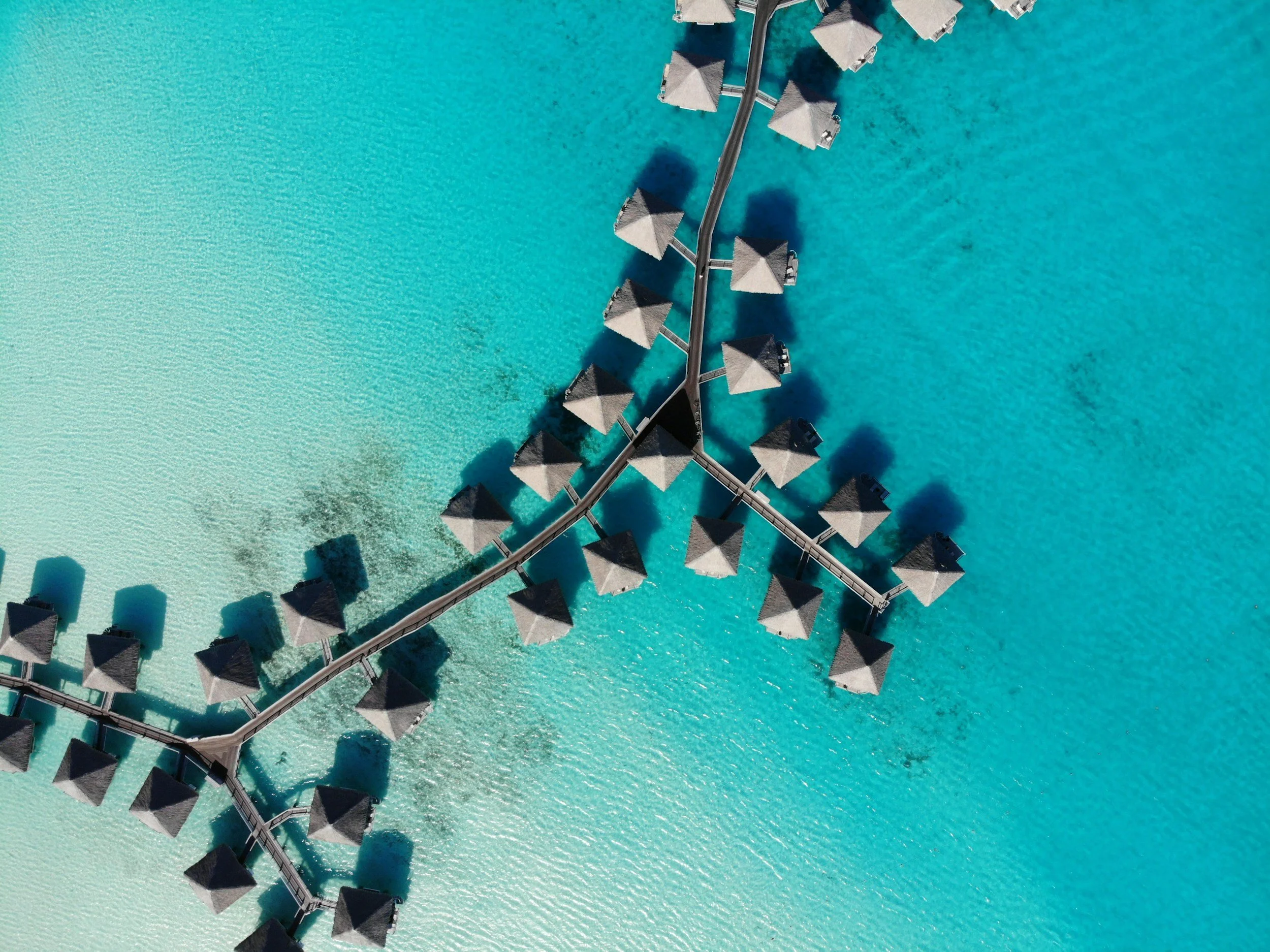 An aerial view of overwater bungalows with thatched roofs in turquoise ocean water.