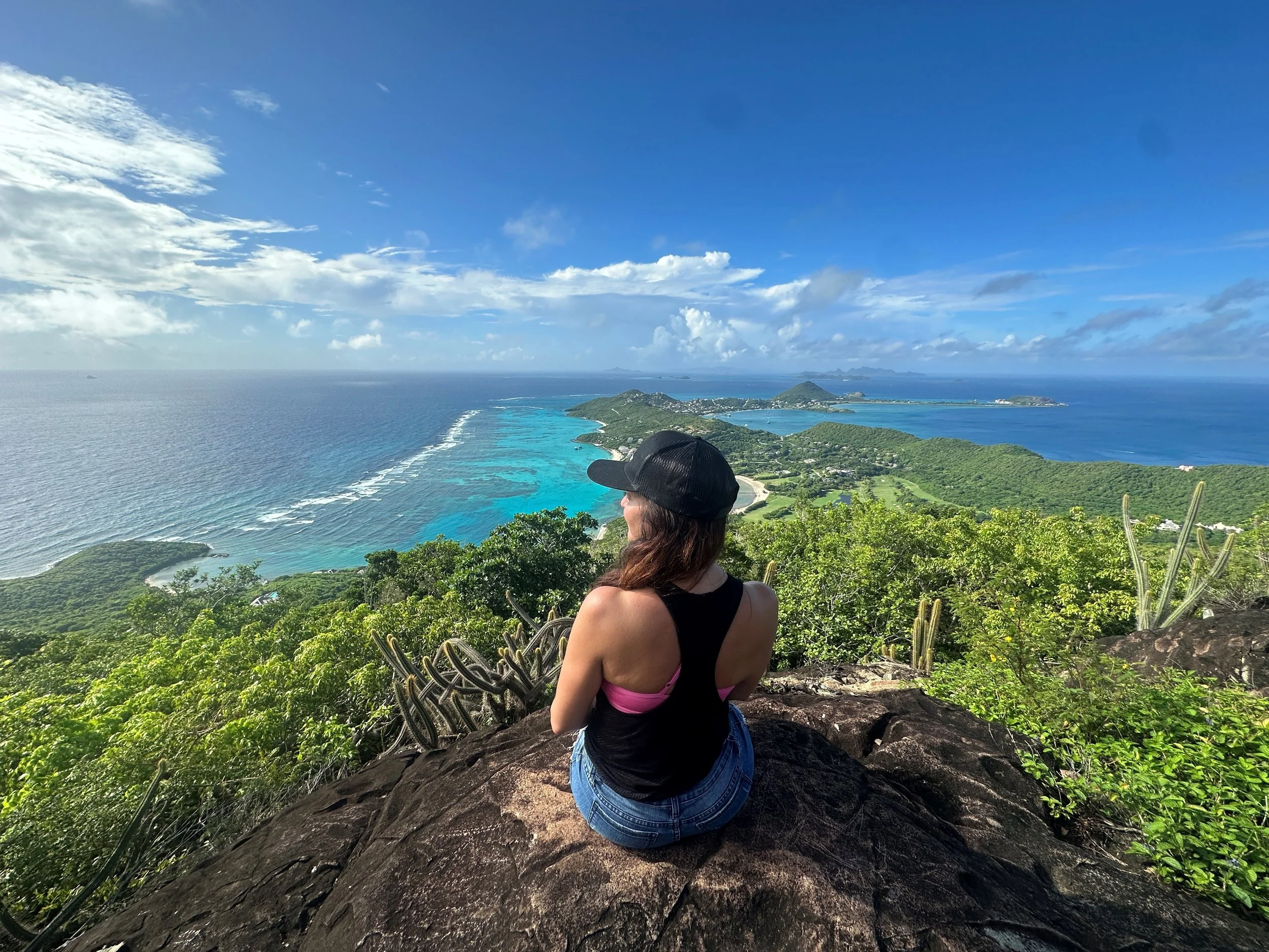 A woman sitting on a rock overlooking a scenic tropical coastline with turquoise waters, green islands, and a blue sky with clouds.