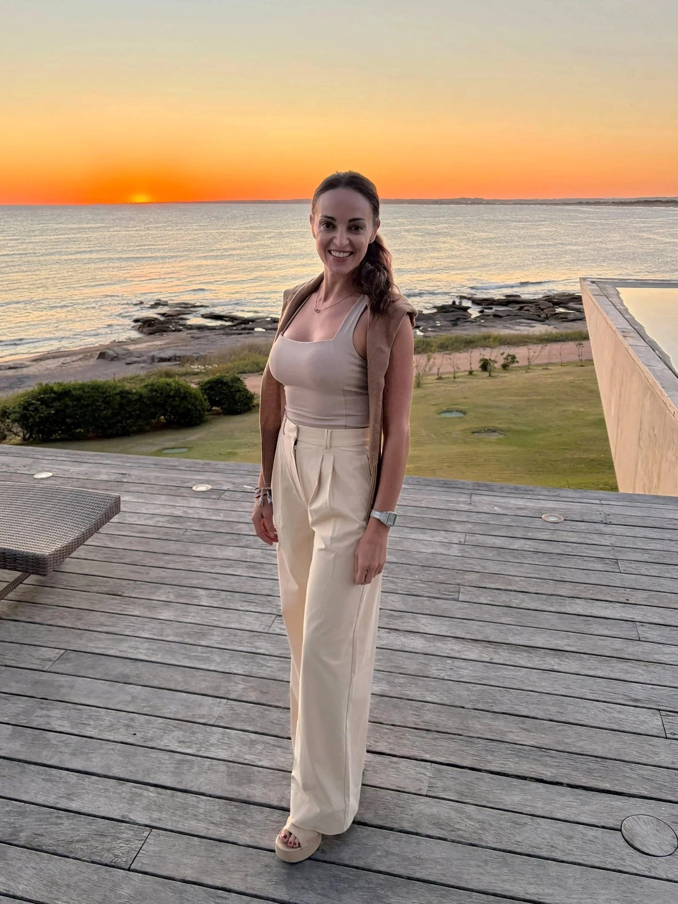 A woman standing on a wooden deck near the coast during sunset, wearing a beige top, cream pants, and a light brown sleeveless jacket, smiling at the camera with the ocean and sunset in the background.