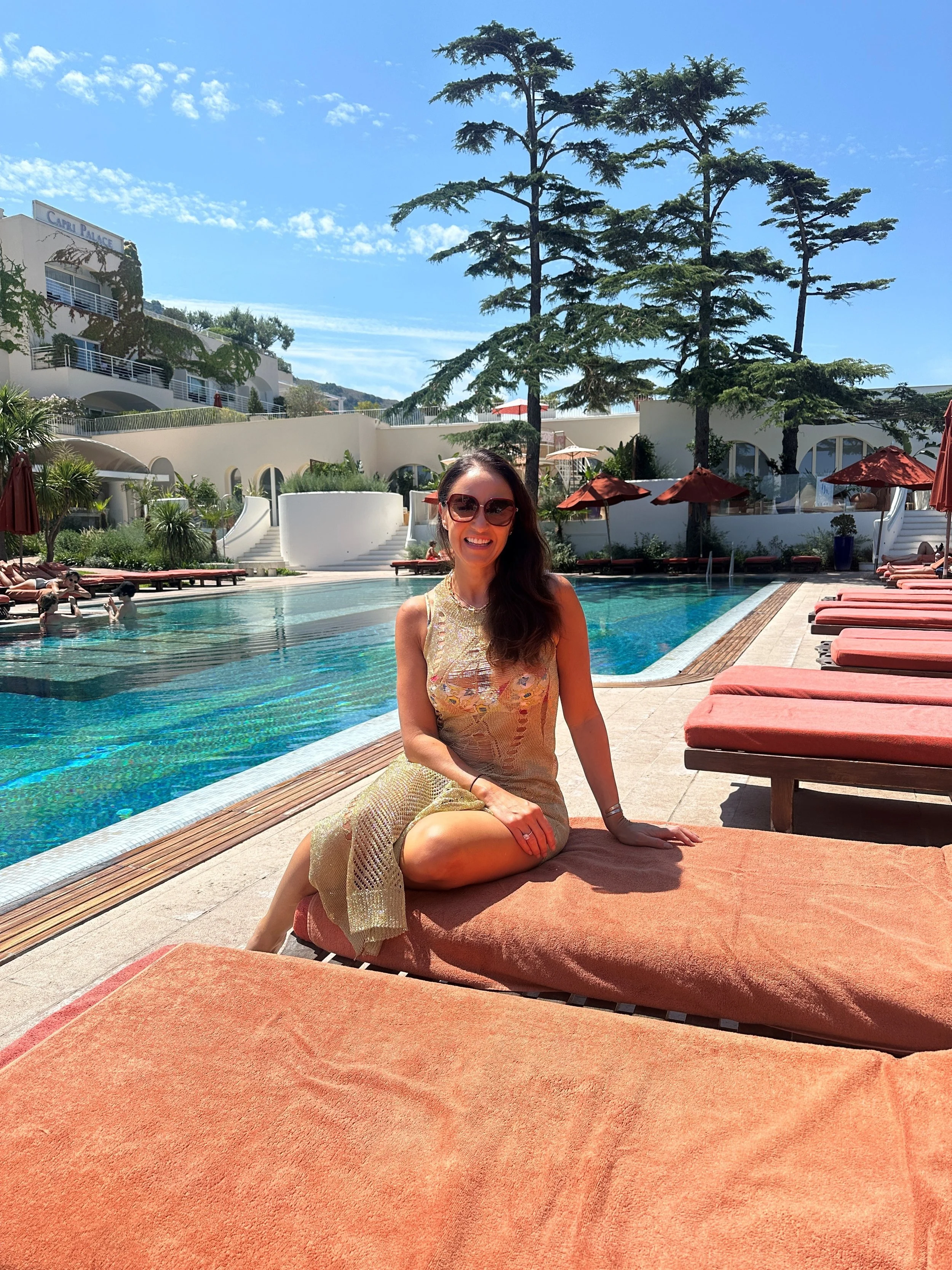 A woman with long, wavy brown hair and sunglasses, wearing a sleeveless beige dress, sitting on an orange poolside lounge chair near a swimming pool at a resort with white buildings, tall trees, and umbrellas.