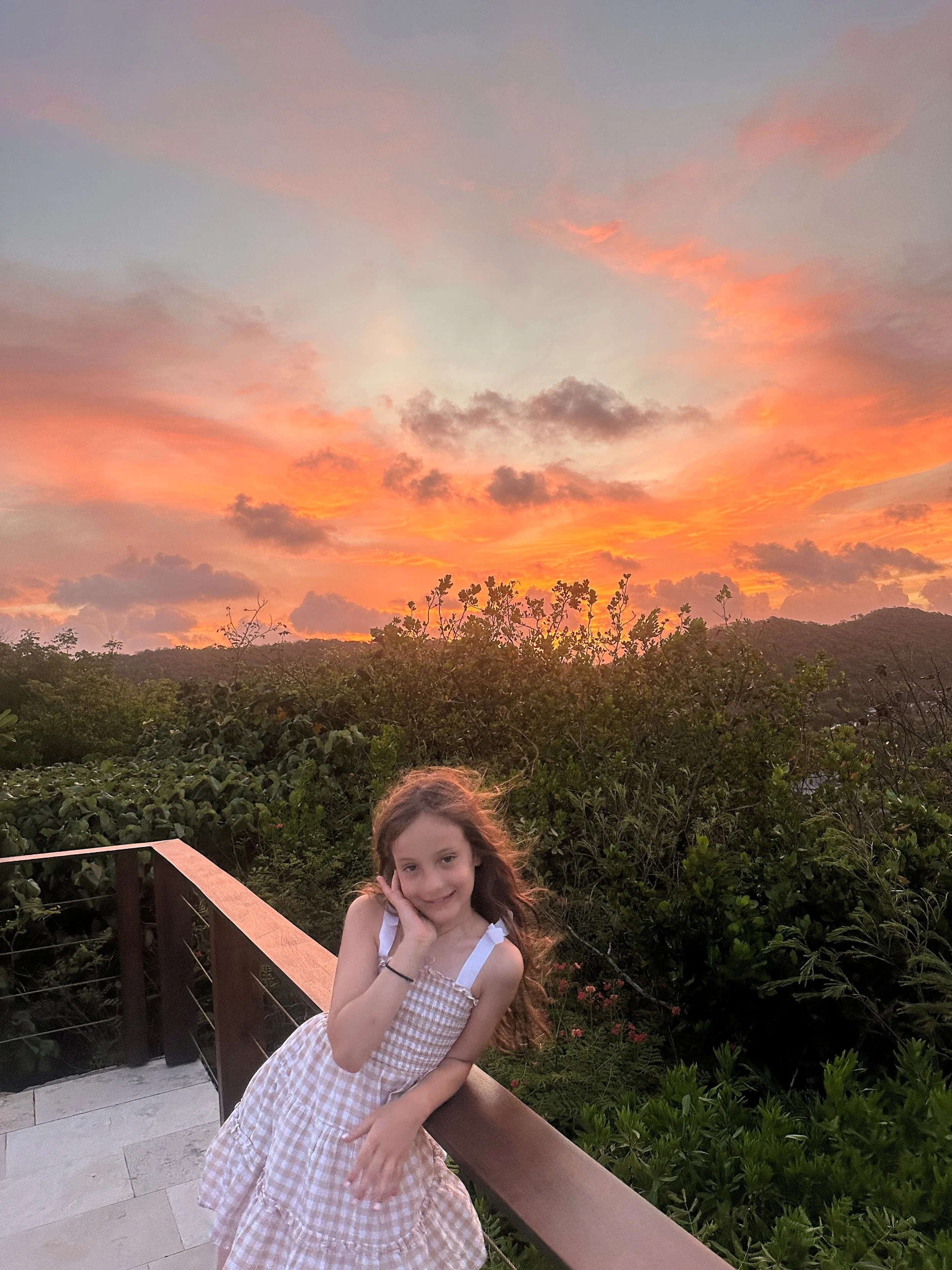 A young girl with long hair wearing a beige checkered dress, leaning on a wooden railing on a balcony during a sunset with a sky filled with orange, pink, and purple clouds, and surrounded by lush green vegetation.