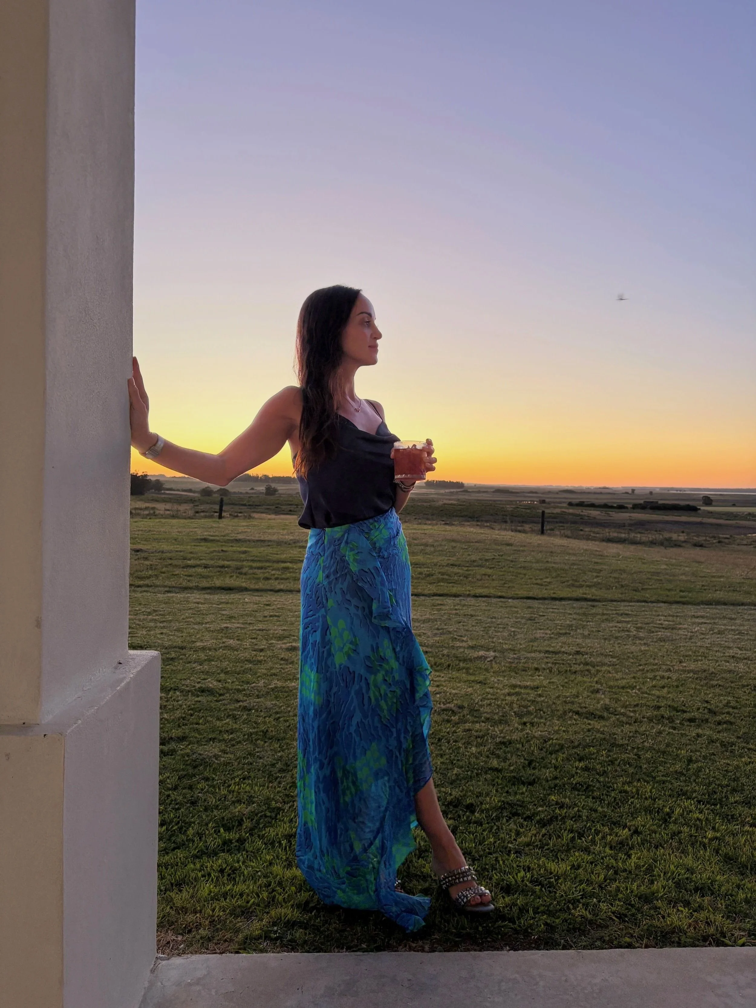 A woman in a black top and blue patterned skirt standing outside at sunset, holding a drink in one hand, with her other hand resting on a wall, looking gracefully into the distance.