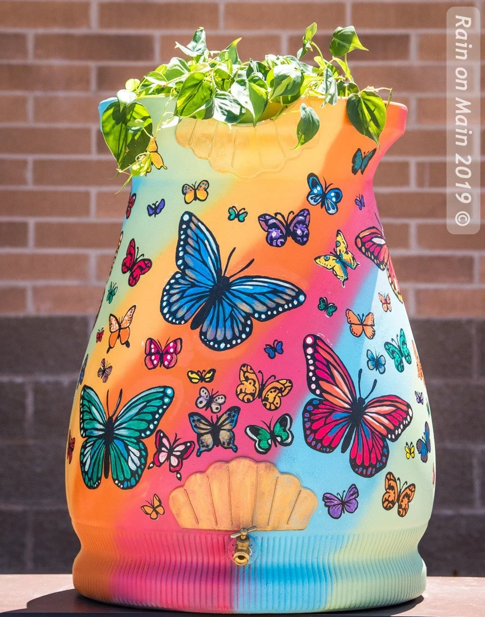 Colorful rain barrel with butterfly and floral pattern, on a flat surface against a brick wall background.