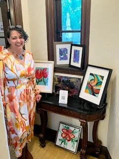 Aimee Bruno, Artist standing next to a display of framed colorful artwork on a dark wooden table inside a room with a window.