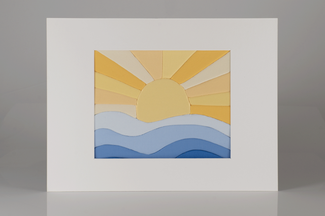 A fabric art piece of a sunrise over blue waves, framed with white matting.