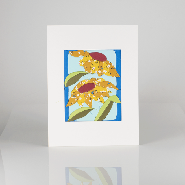 A framed fabric art piece with a colorful fabrics of three yellow flowers with orange centers and white accents, surrounded by green leaves, on a blue background.