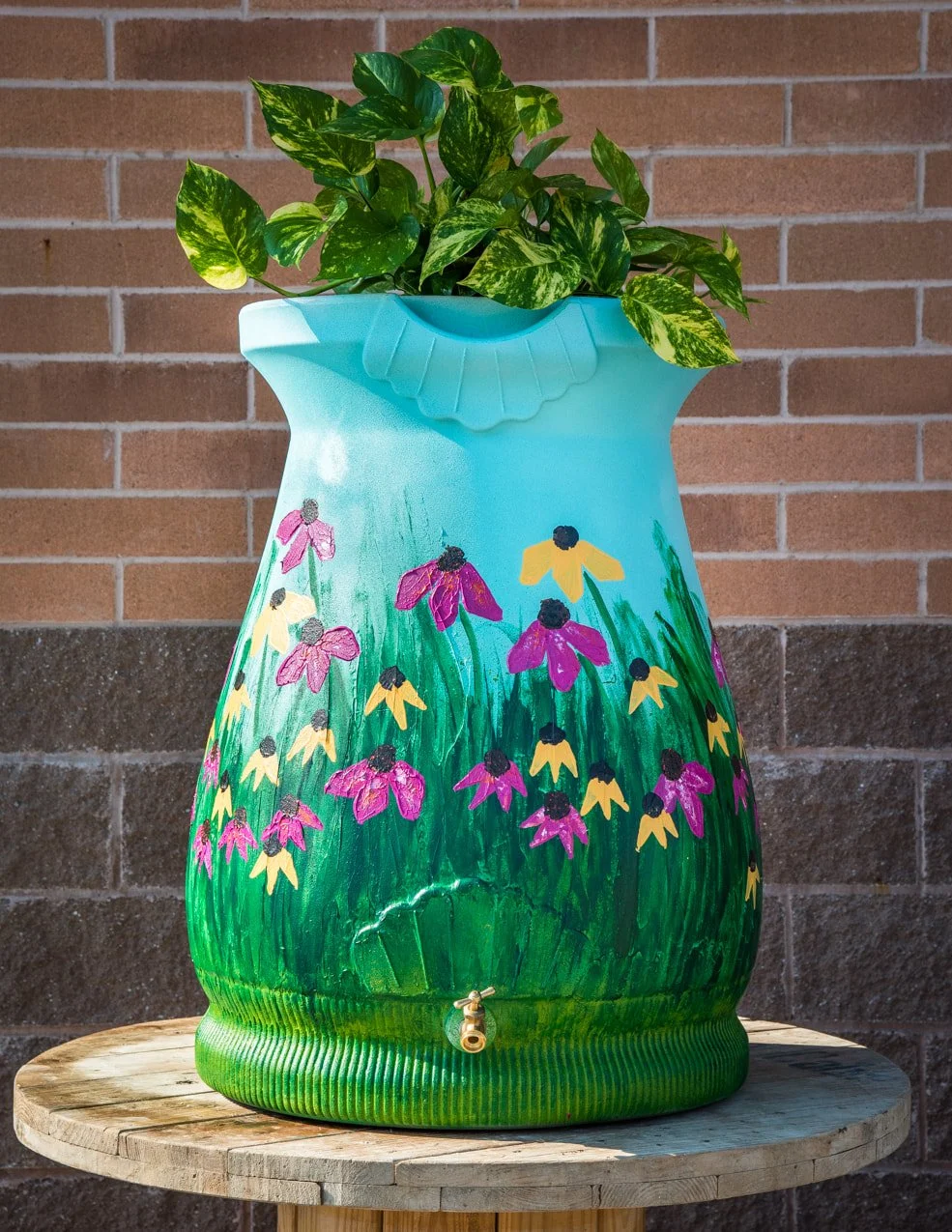 Decorative barrel painted with pink and yellow flowers, placed on a wooden table against a brick wall, with a green leafy plant on top.