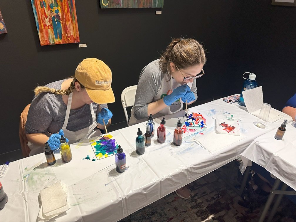 Two women are sitting at a table covered with art supplies, painting. 