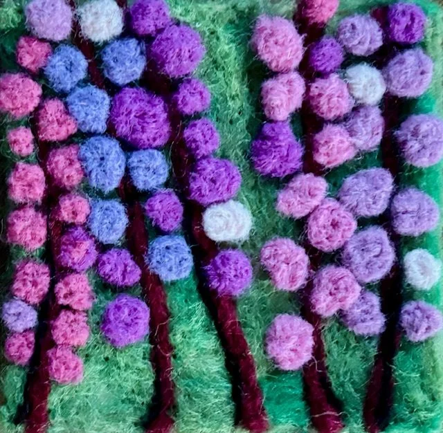 Colorful floral needle felted painting featuring pink and purple pom-pom flowers with green stems on green fabric background.