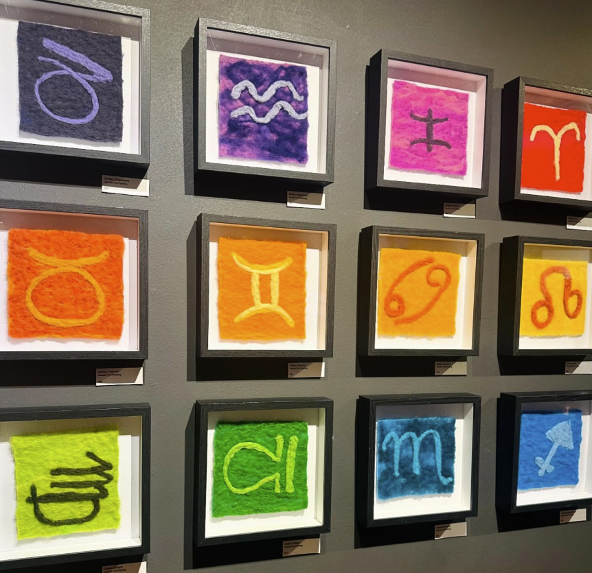 A wall display featuring twelve framed artworks, each with a colorful background and a zodiac sign painted in a contrasting color. The artworks include representations of Aries, Taurus, Gemini, Cancer, Leo, Virgo, Libra, Scorpio, Sagittarius, Capricorn, Aquarius, and Pisces.