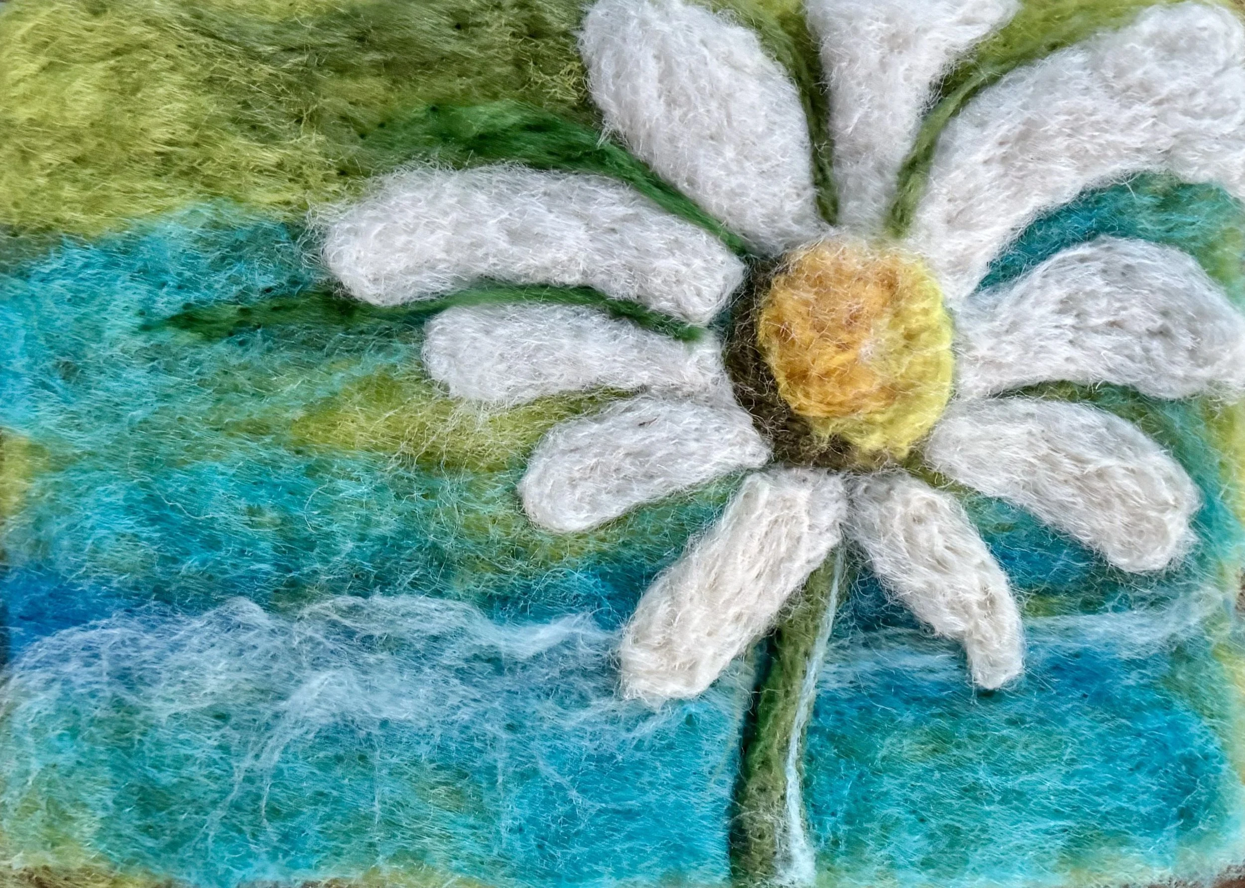 A needle-felted art piece depicting a daisy flower with white petals, a yellow center, green stem, against a background of blue sky and green grass.