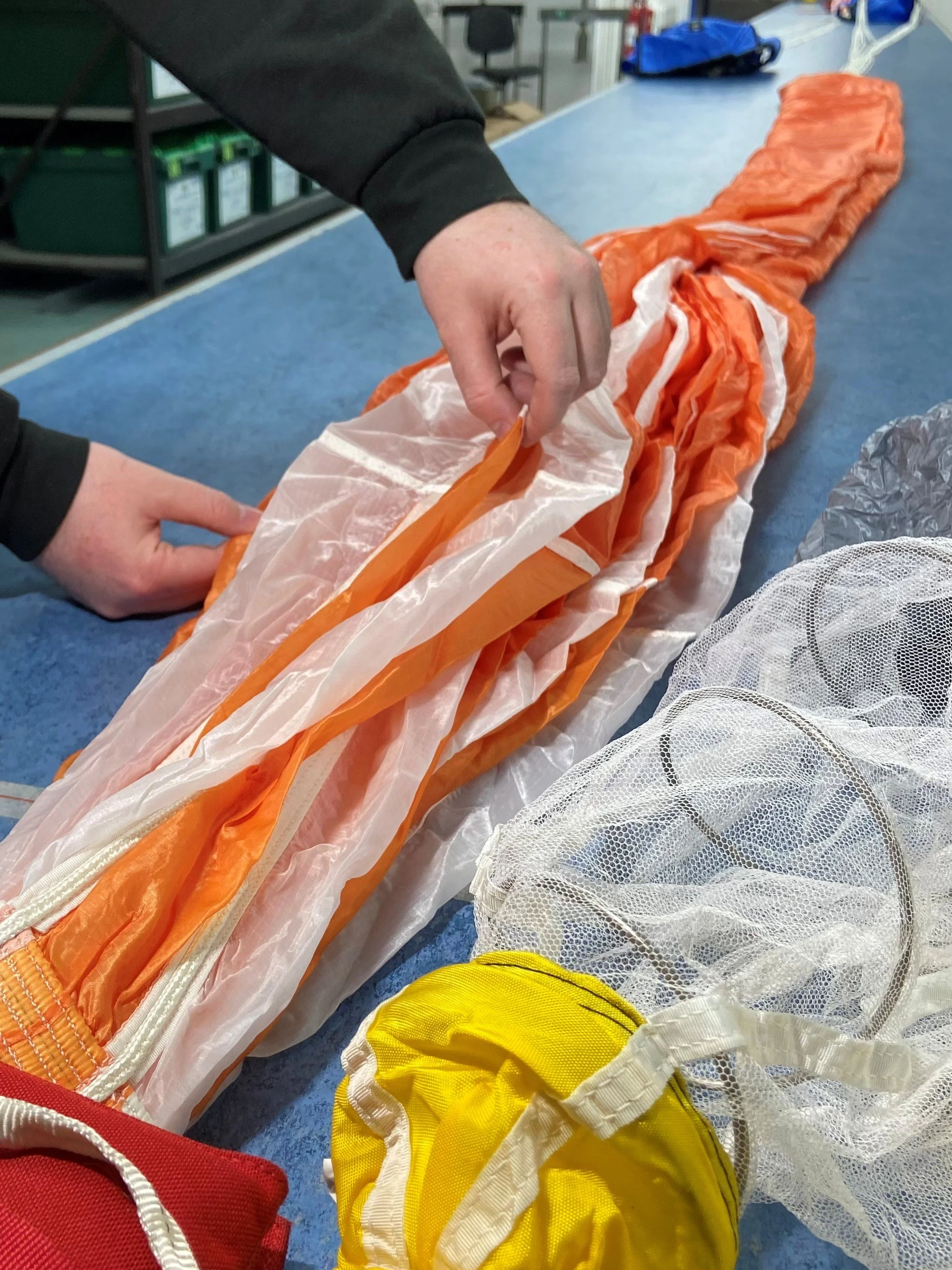 Person packing or preparing an orange and white parachute on a blue table, with other yellow and white gear nearby.