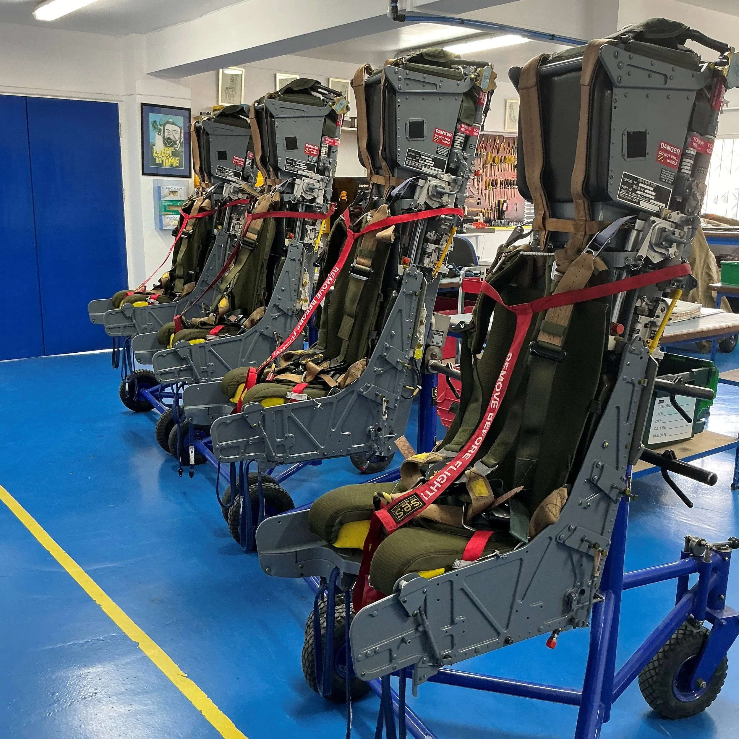 Four aircraft ejection seats lined up in a workshop with blue flooring and various tools in the background.