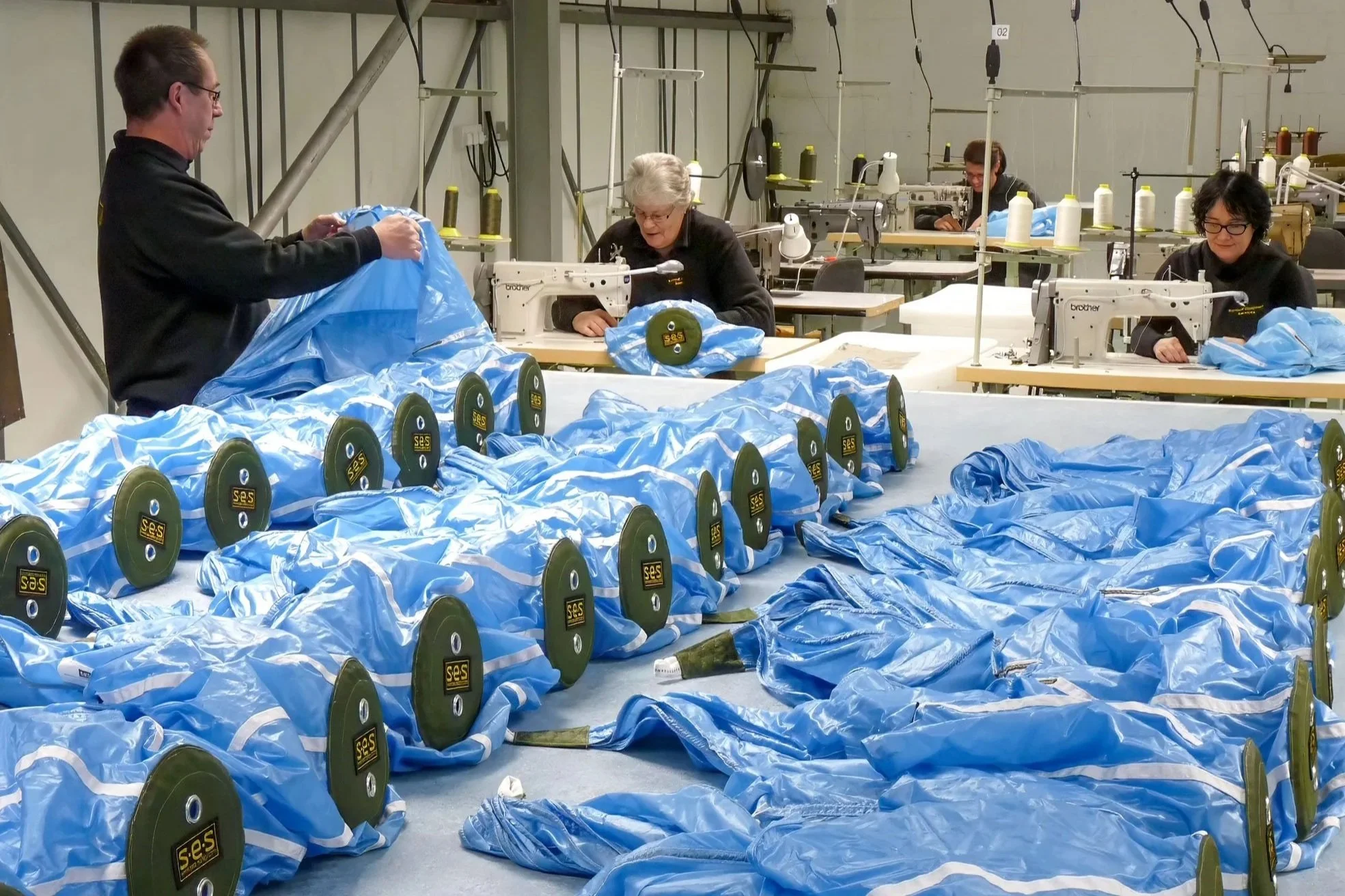 Workers in a factory sewing blue drogue parachutes at sewing machines, with completed parachutes laid out on the table.
