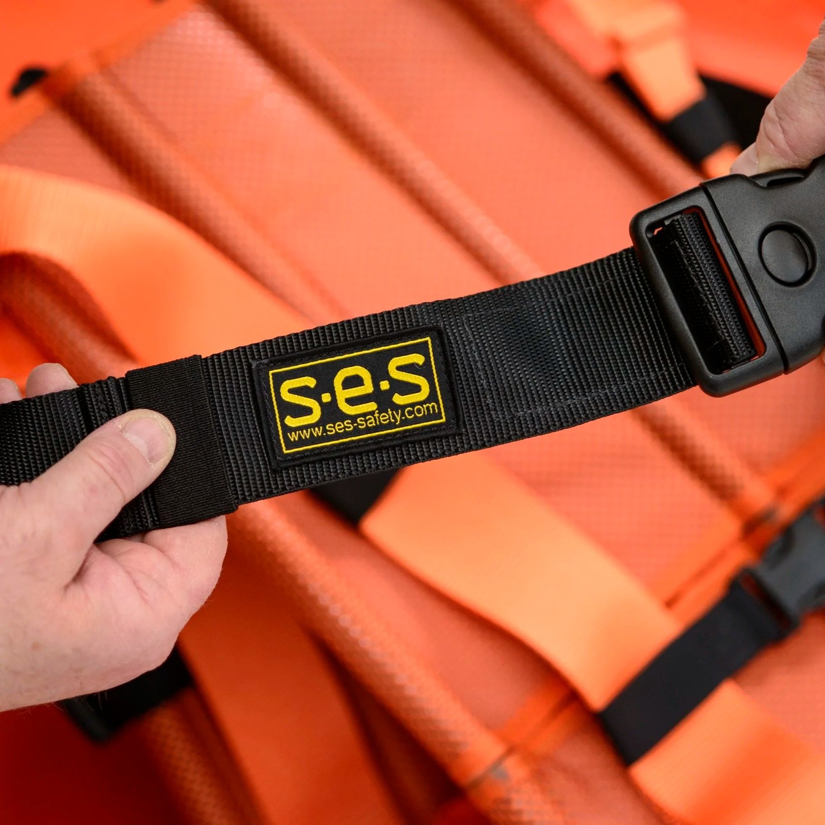 Close-up of a black safety strap labeled 'S.E.S' with a yellow logo and website, held in hand, with an orange rescue backboard in the background.