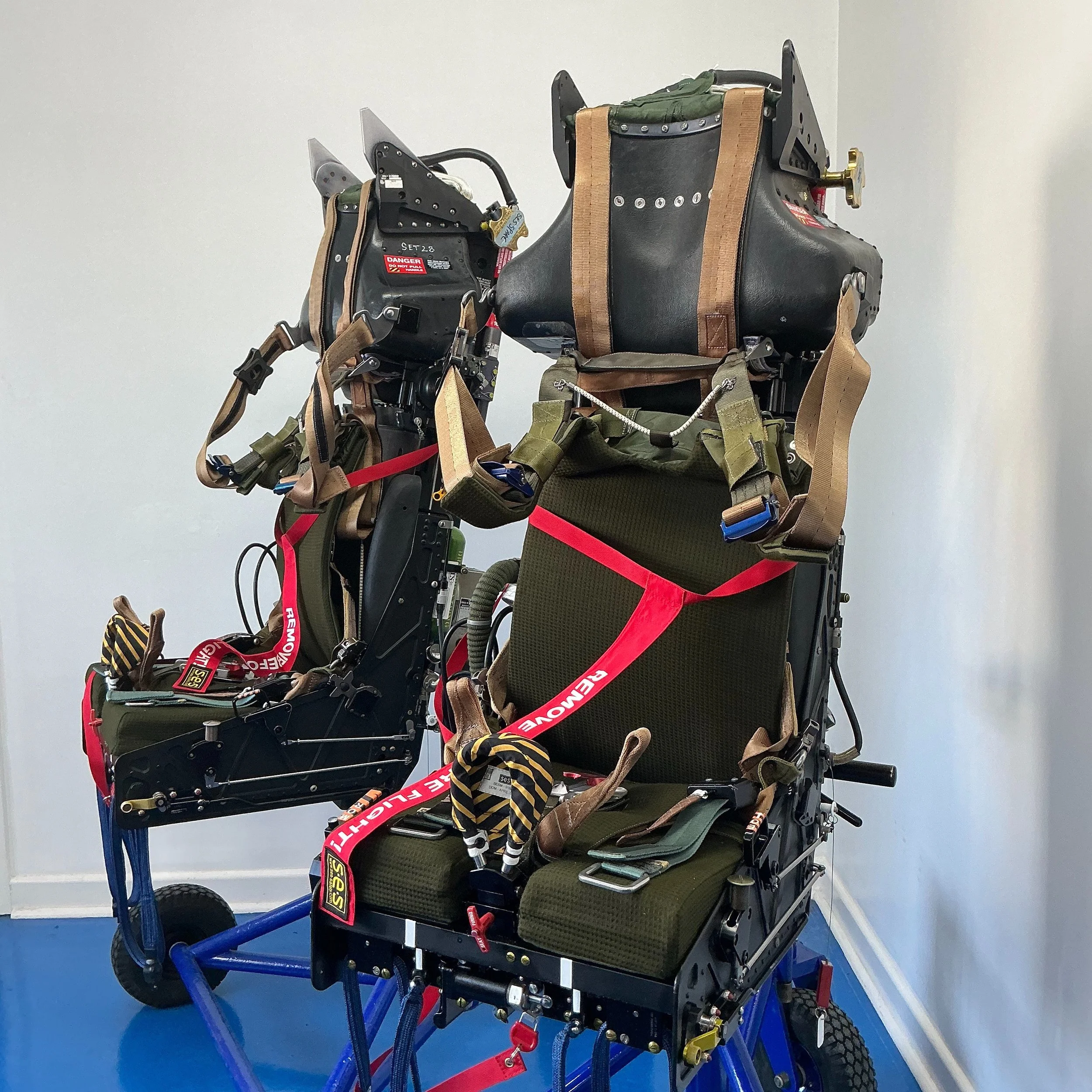 Aviation ejection seat with harnesses, padding, and electronic components, mounted on a blue frame with wheels.