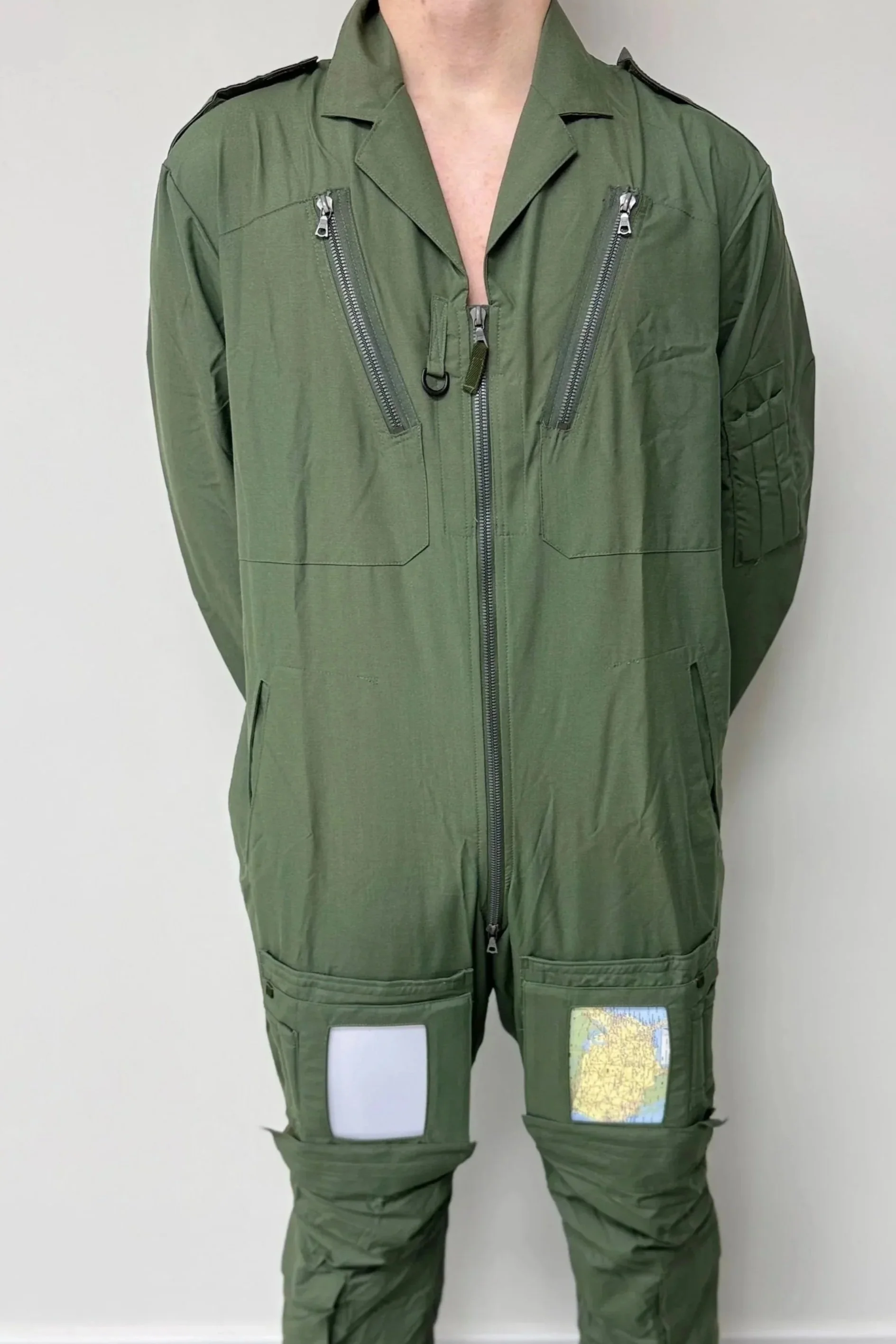 A person wearing a green tactical or military jumpsuit with multiple zippered pockets and patches, standing against a plain wall.