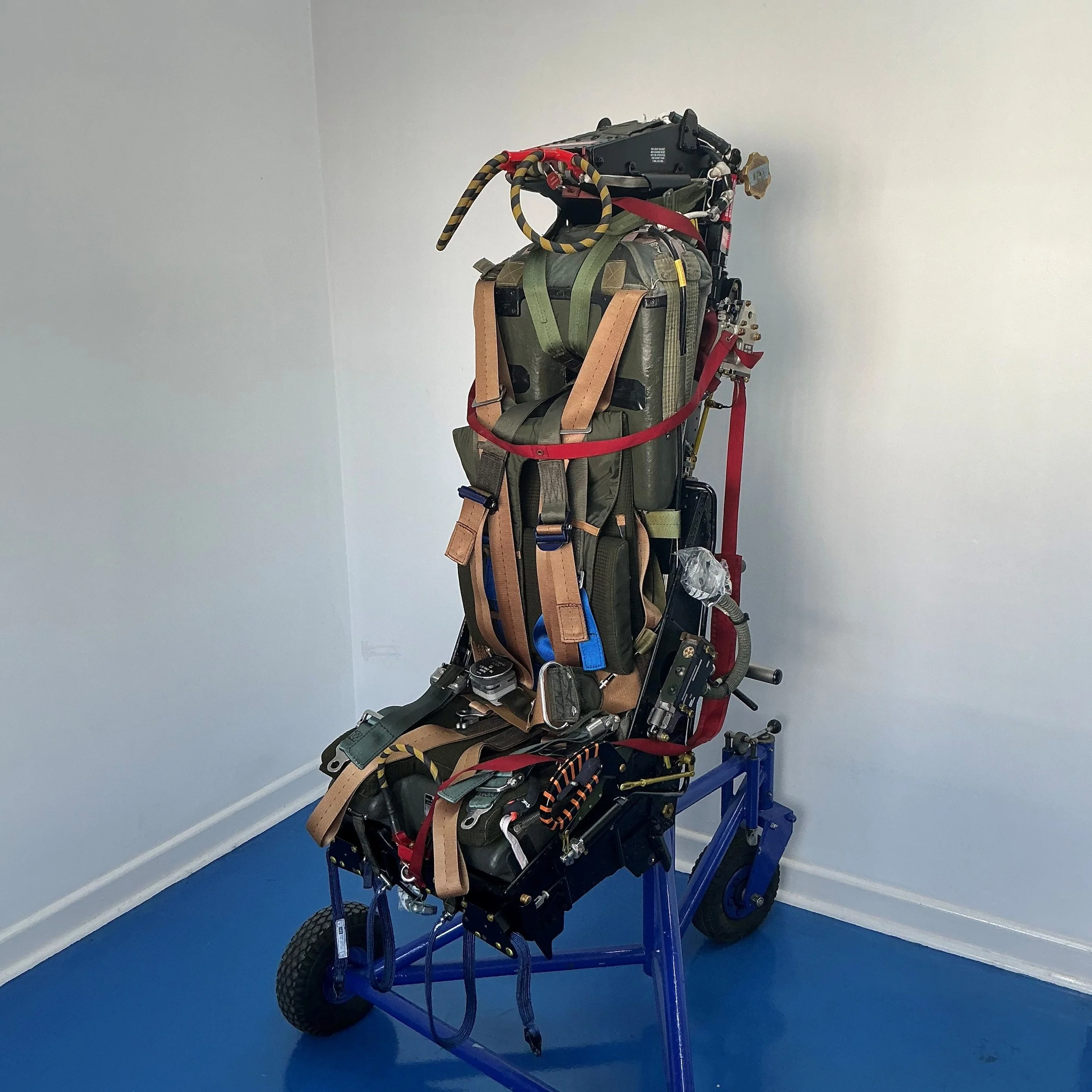 An aircraft ejection seat, mounted on a blue wheeled frame, positioned against a corner of a white room with a blue floor.