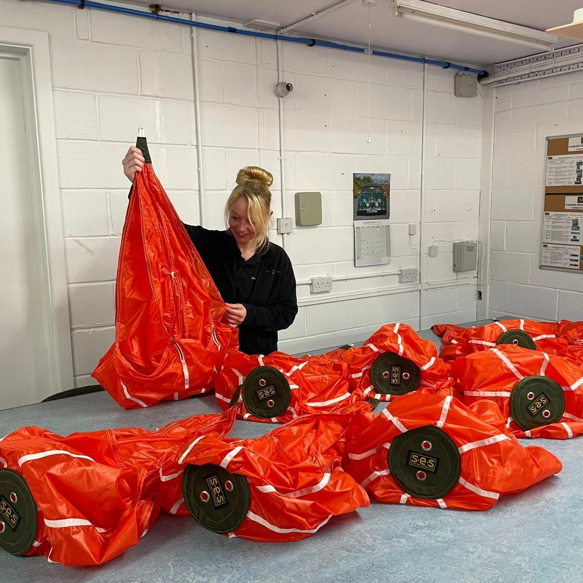 A woman is smiling as she handles a bright orange drogue parachute. The parachutes have black circular bases with labels that read 'S.E.S'. The room has white brick walls with cables, a calendar, and electrical boxes on the wall.