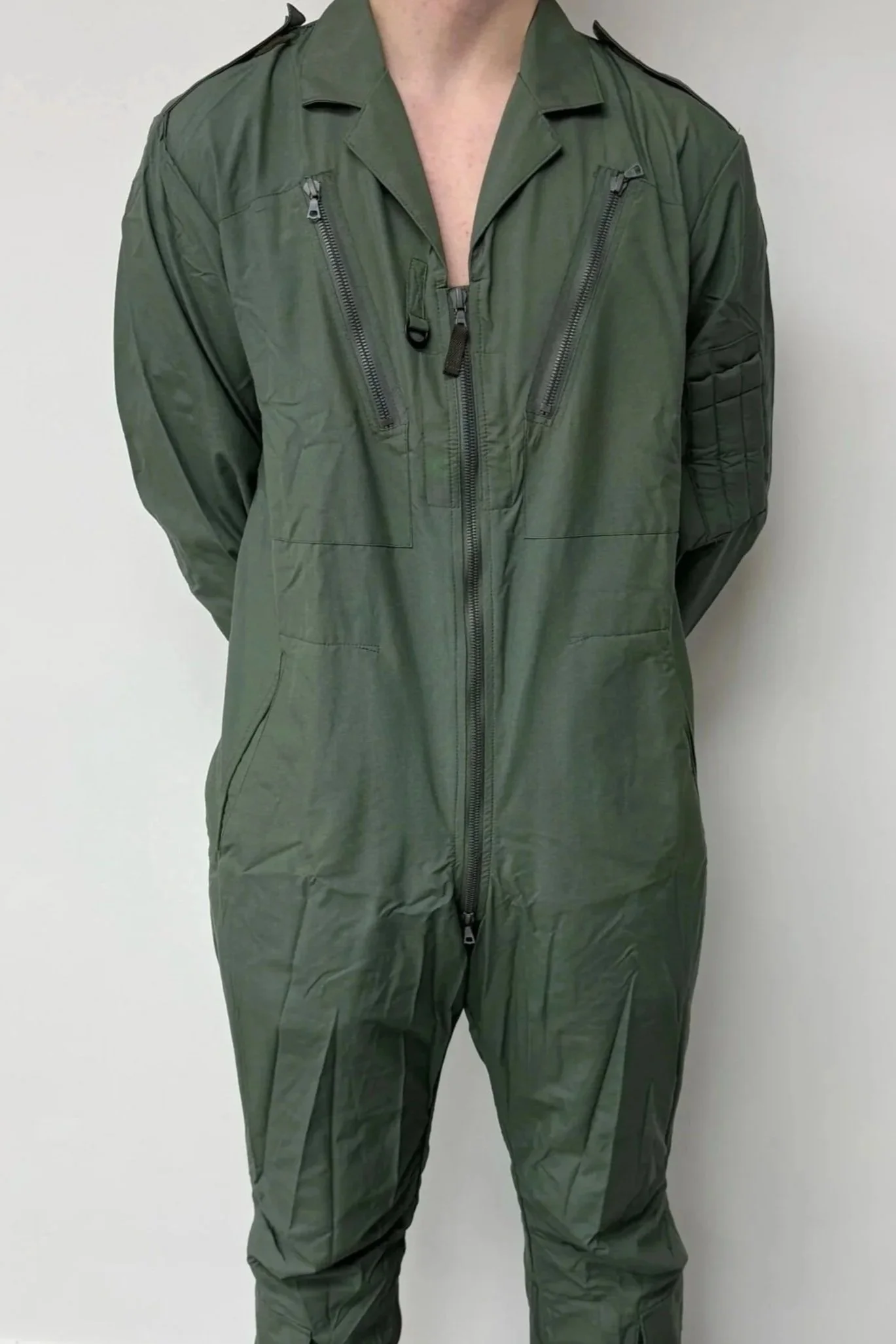 Person wearing a green military-style jumpsuit with a front zipper and multiple zippered pockets, standing against a plain wall.
