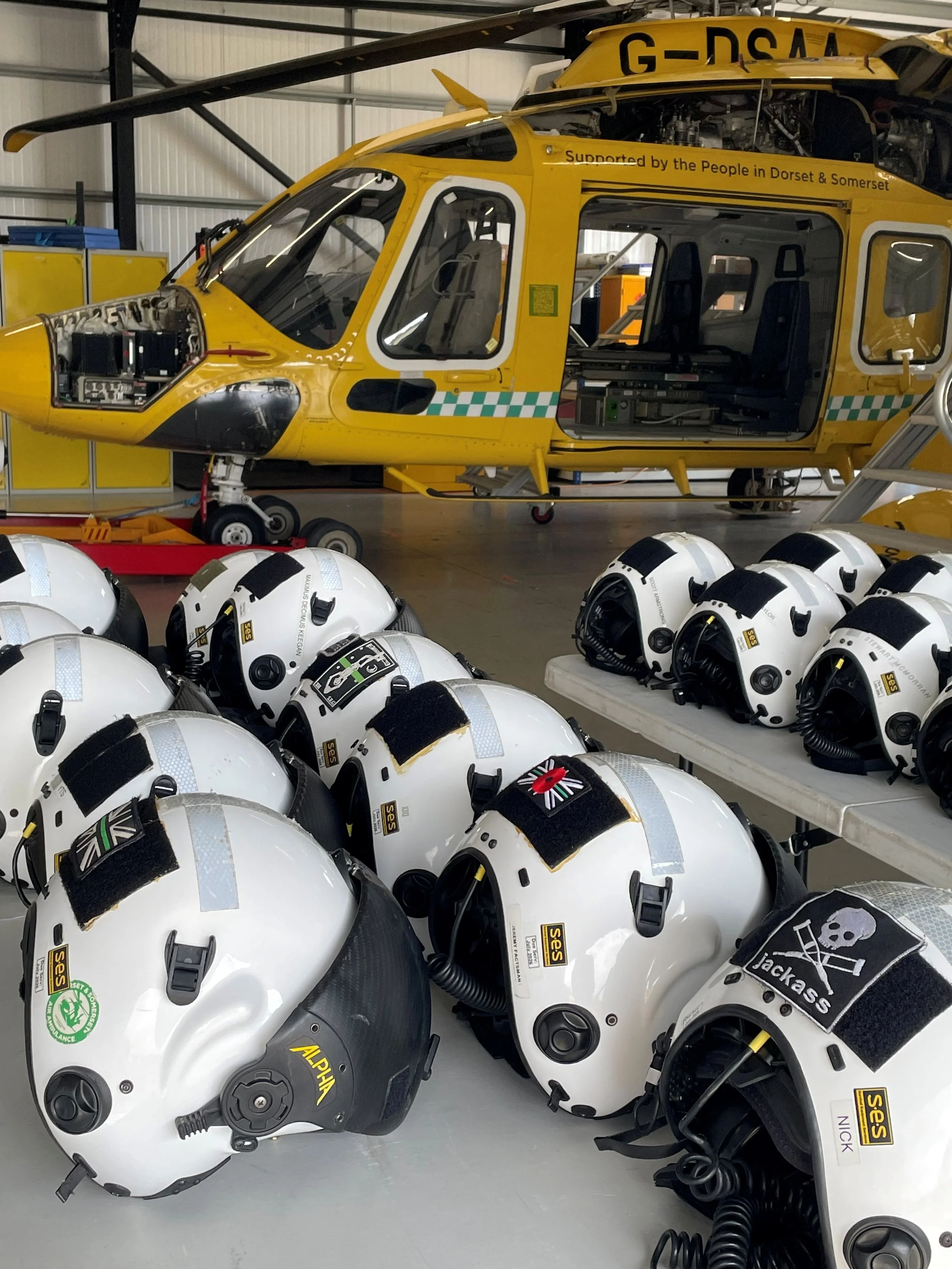 Multiple white helicopter helmets with microphone cords on a table and a yellow rescue helicopter in the hangar behind them.