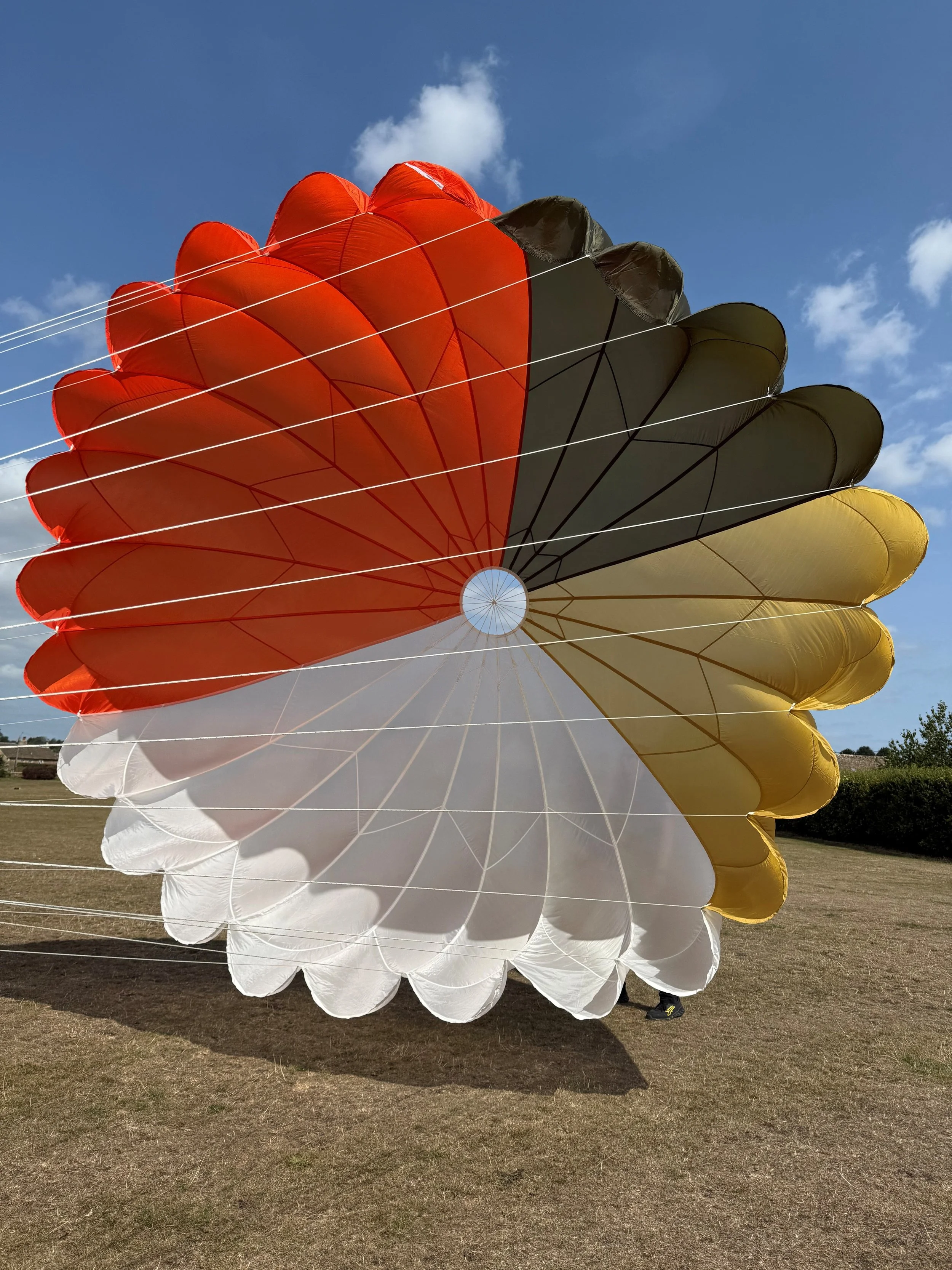 A multicolored parachute with segments in red, black, yellow, and white, partially inflated on a grassy field against a blue sky with clouds.