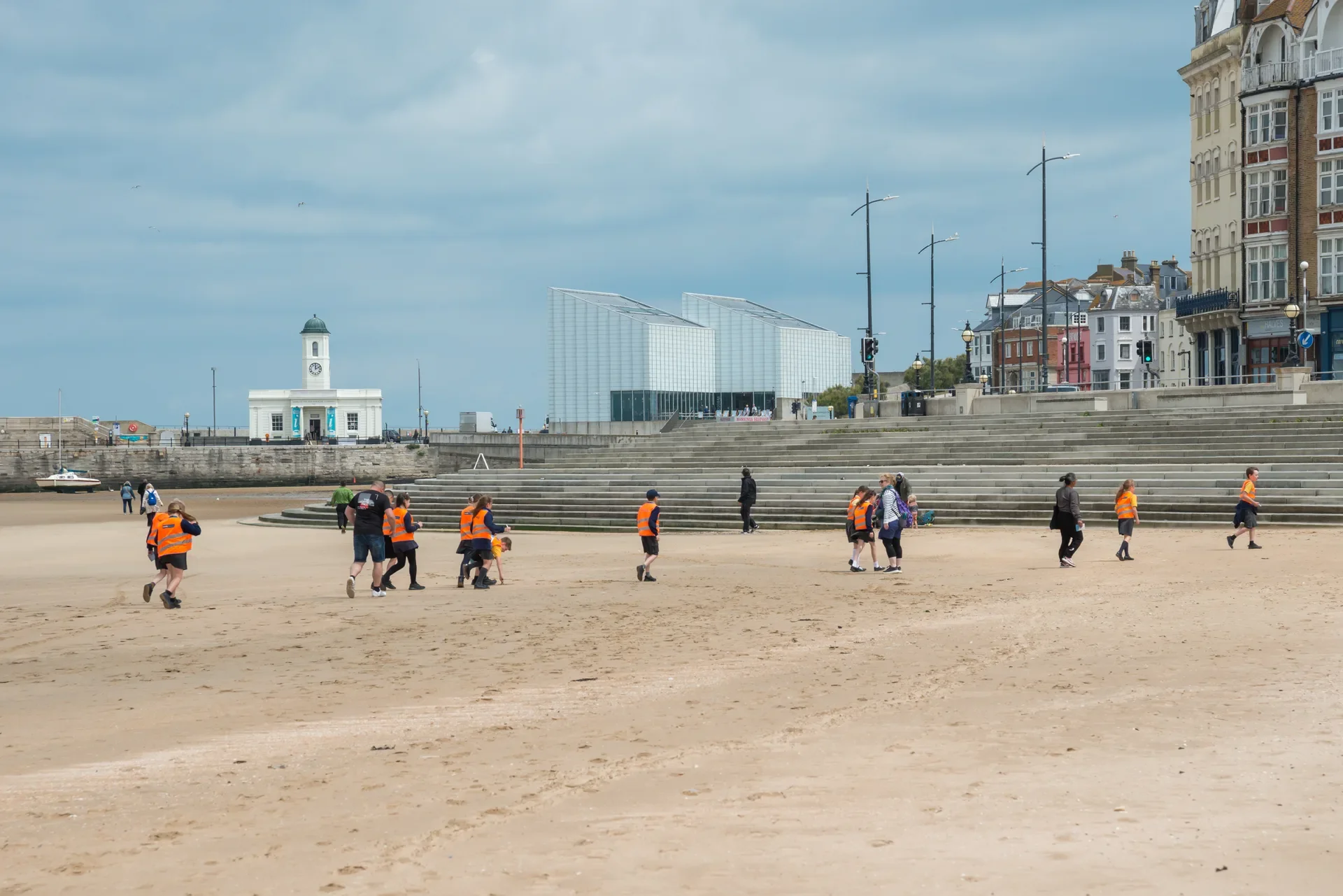Group of children and adults wearing orange safety vests walking on a Margate beach with the Turner Contemporary, Visitor Information Centre and the Margate Steps in the background.