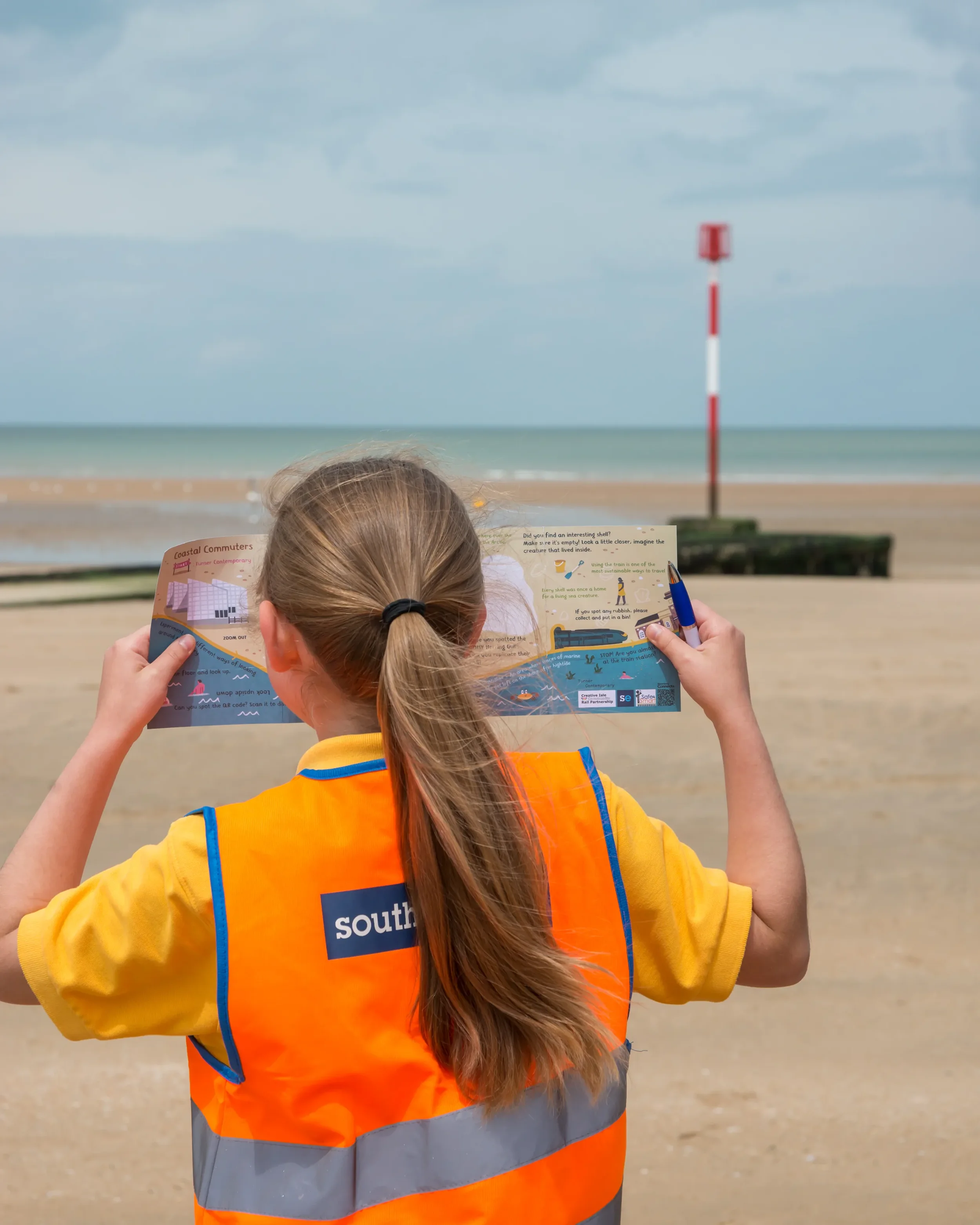 A young girl in a Southeastern hi-vis vest reads a colorful 'coastal commuters' resource by Creative Isle Community Rail Partnership on Margate main sands beach.