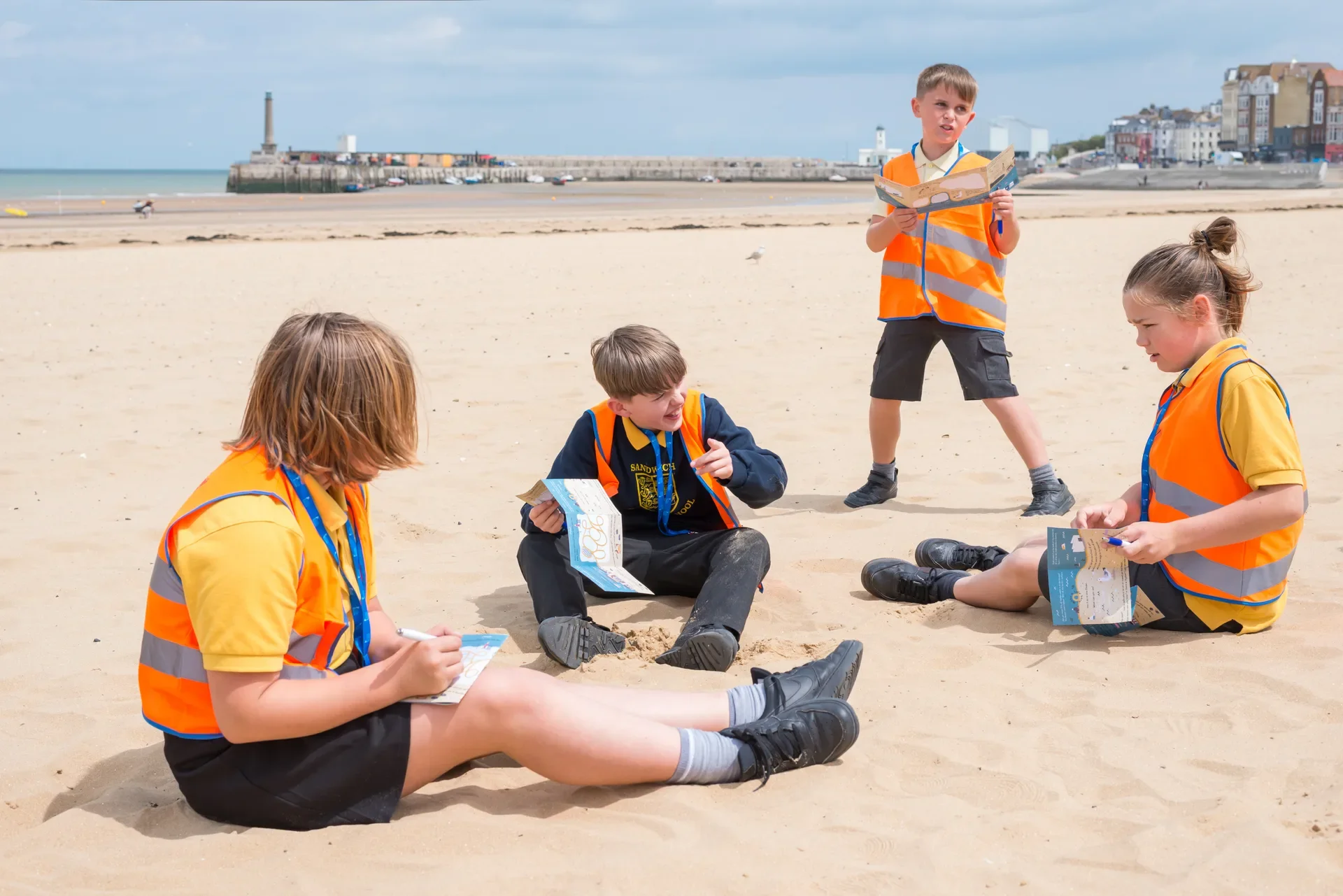 Four children wearing reflective Southeastern Railway branded hi-vis vests sitting and standing on Margate beach, holding maps and writing, with Margate Harbour Arm and the Turner Contemporary in the background.