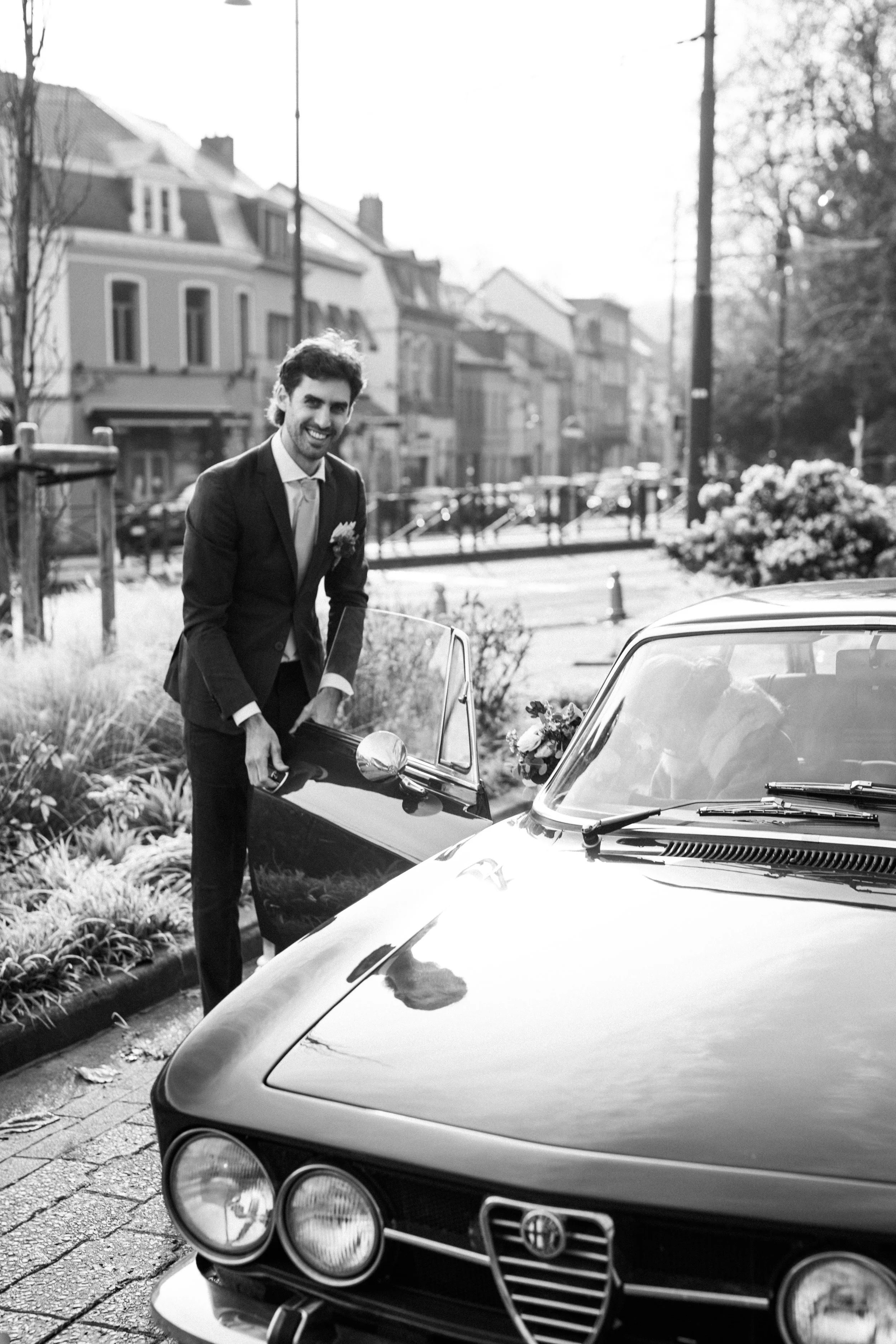 Un homme habillé en costume de mariage, souriant, sort d'une voiture de collection noire sur une rue résidentielle, avec des maisons et des arbres en arrière-plan.