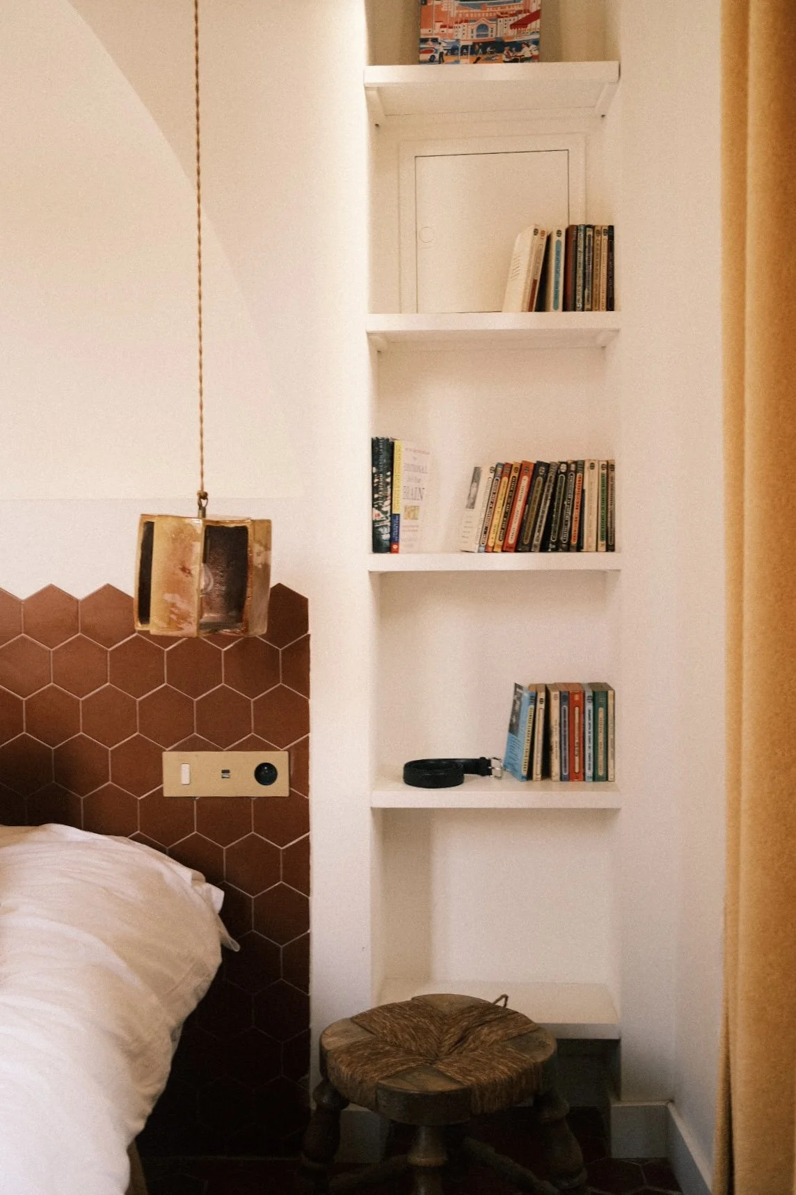 Une étagère blanche avec des livres, un porte-clés, et une petite boîte colorée en haut d'un mur blanc à côté d'un lit avec une tête de lit en carreaux hexagonaux bruns. Il y a aussi un petit tabouret en bois en dessous de l'étagère et un plafond ave