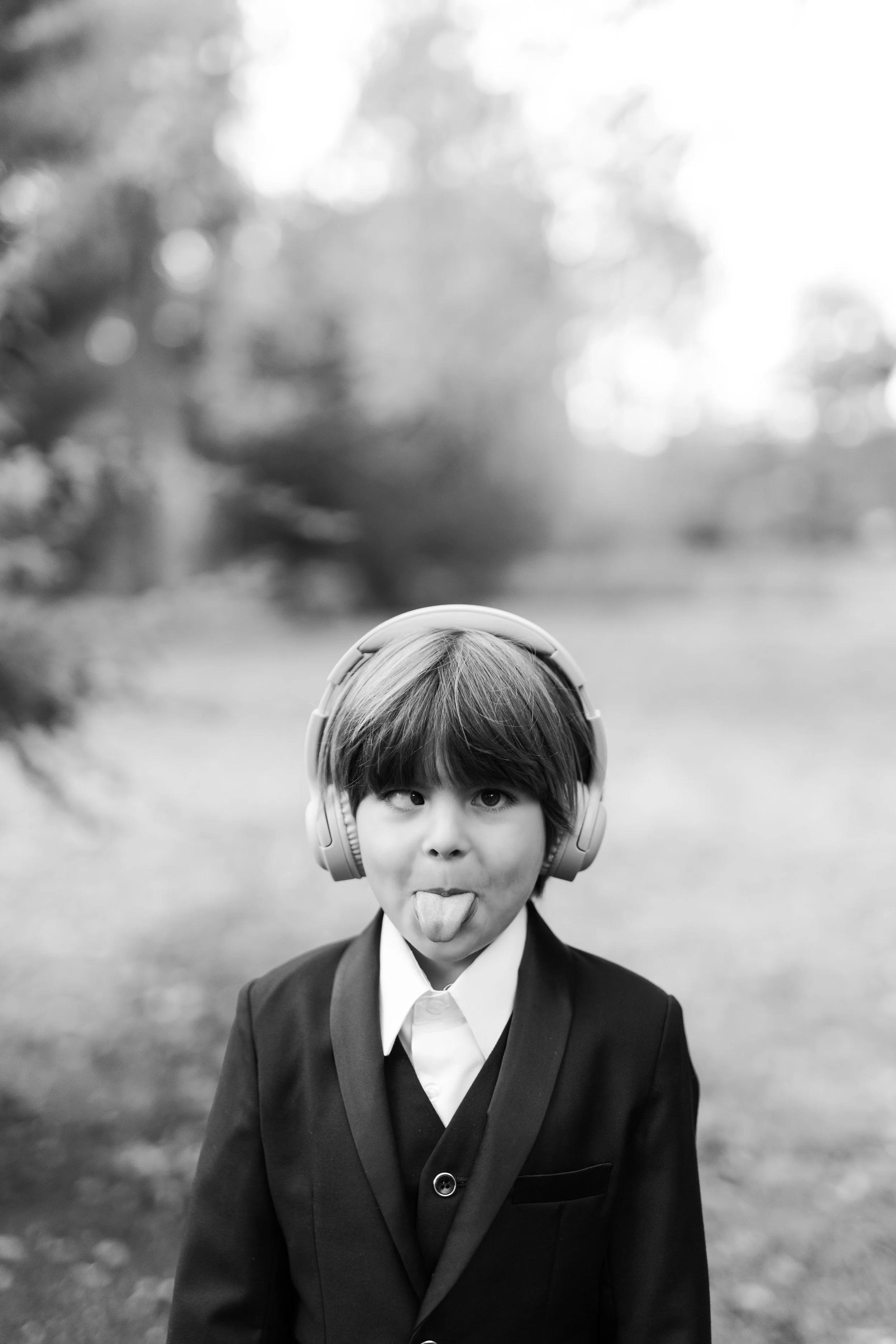 Un garçon en costume noir, blague en tirant la langue avec des écouteurs sur la tête, en extérieur.