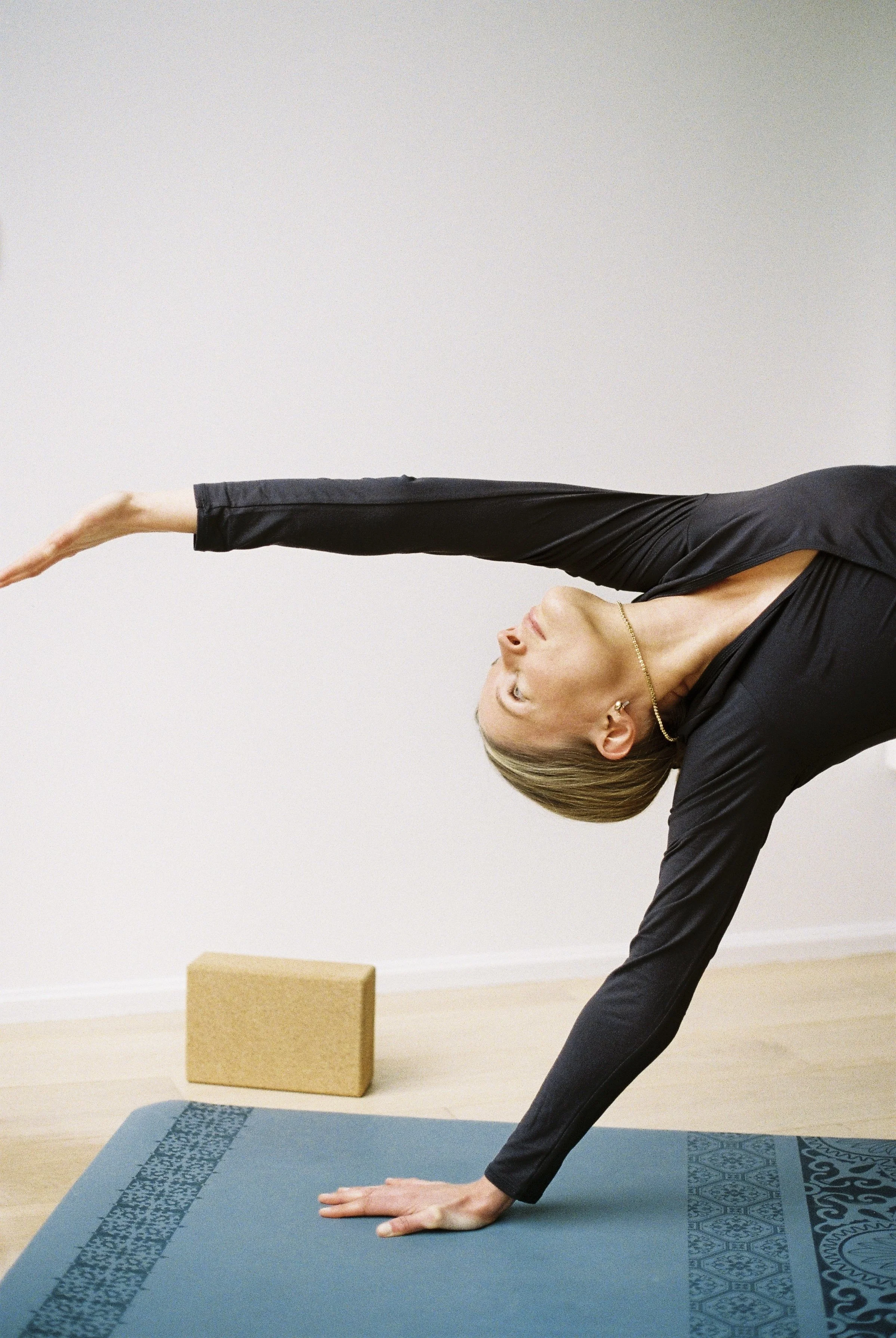Une femme en position de yoga, en équilibre sur une main et un pied, étirant l'autre bras et la tête tournée vers le haut, sur un tapis de yoga dans une pièce