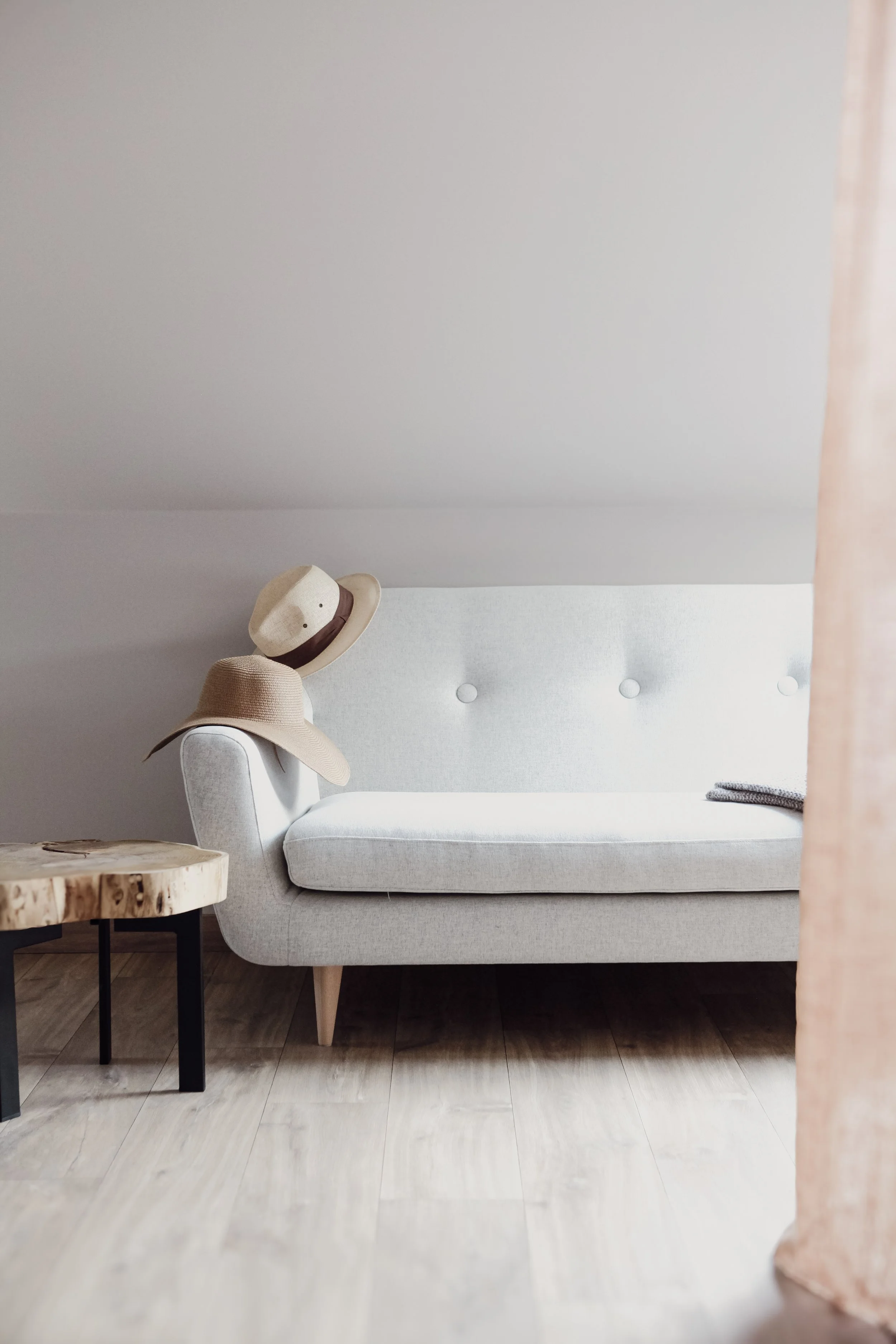 Un canapé gris clair avec deux chapeaux posés dessus, une petite table en bois avec des jambes noires, et un rideau rose pâle à droite.