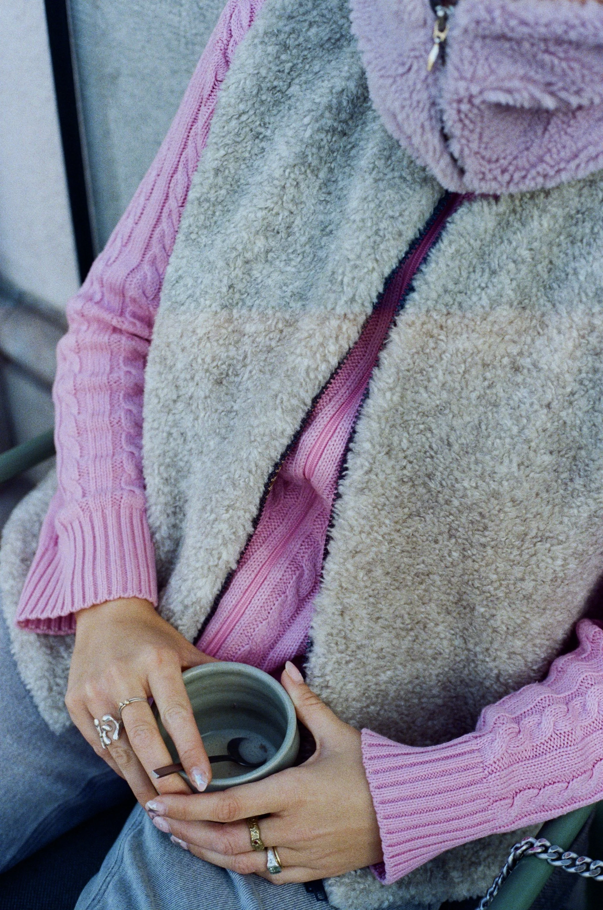 Personne assise tenant une tasse, portant un pull rose à manches longues et un gilet en fausse fourrure beige.