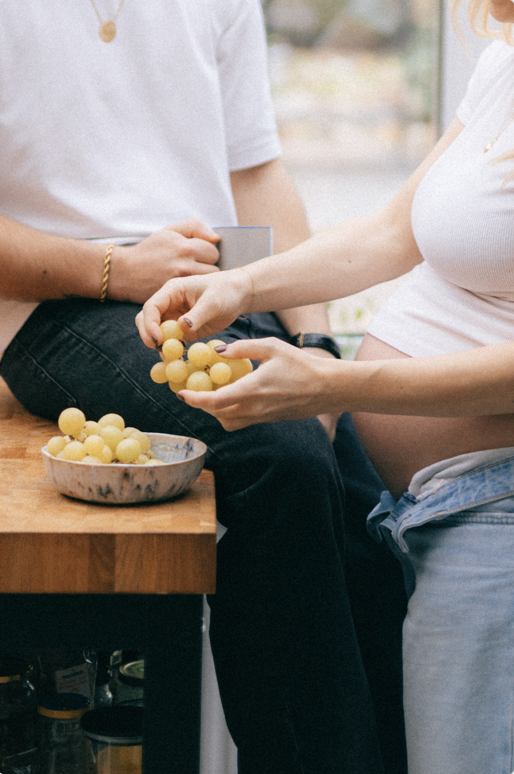 Deux personnes, une assise et une debout, manipulent des grappes de raisins sur un comptoir en bois avec un bol rempli de raisins. La scène se déroule dans un intérieur lumineux.