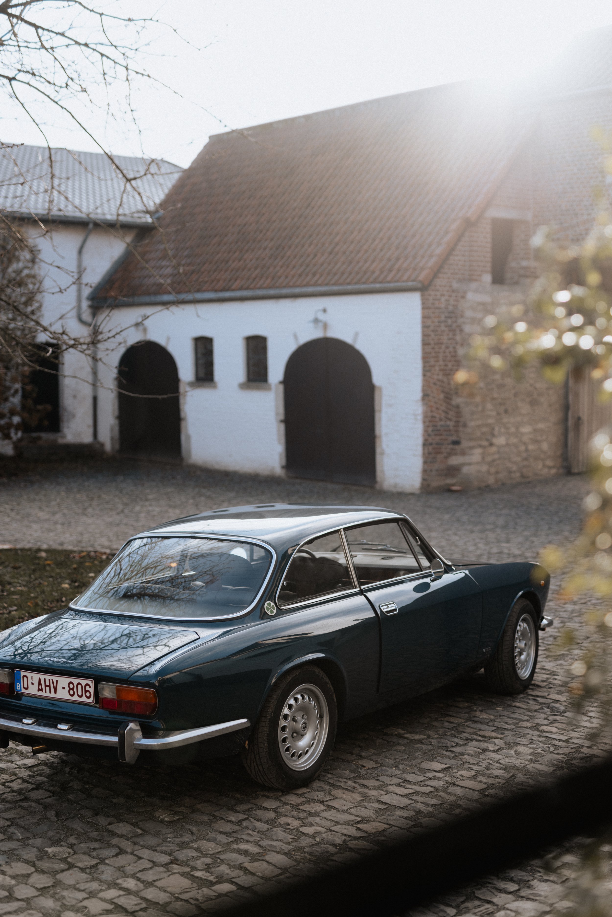 Une voiture ancienne noire stationnée devant une maison avec un mur en pierre et un toit en tuiles, sous une lumière de soleil douce.