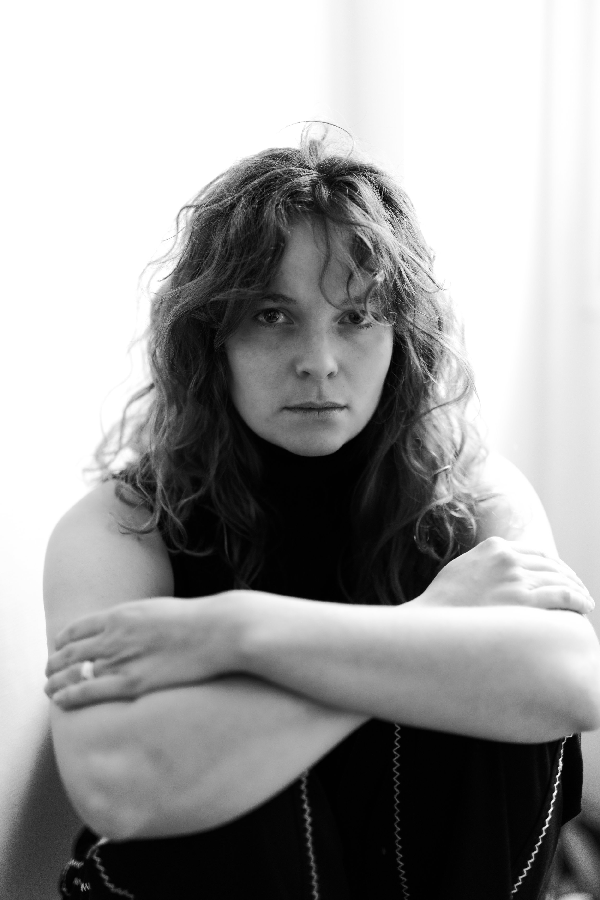 Une femme avec des cheveux bouclés, portant un haut noir, regarde fixement la caméra, les bras croisés sur ses genoux.