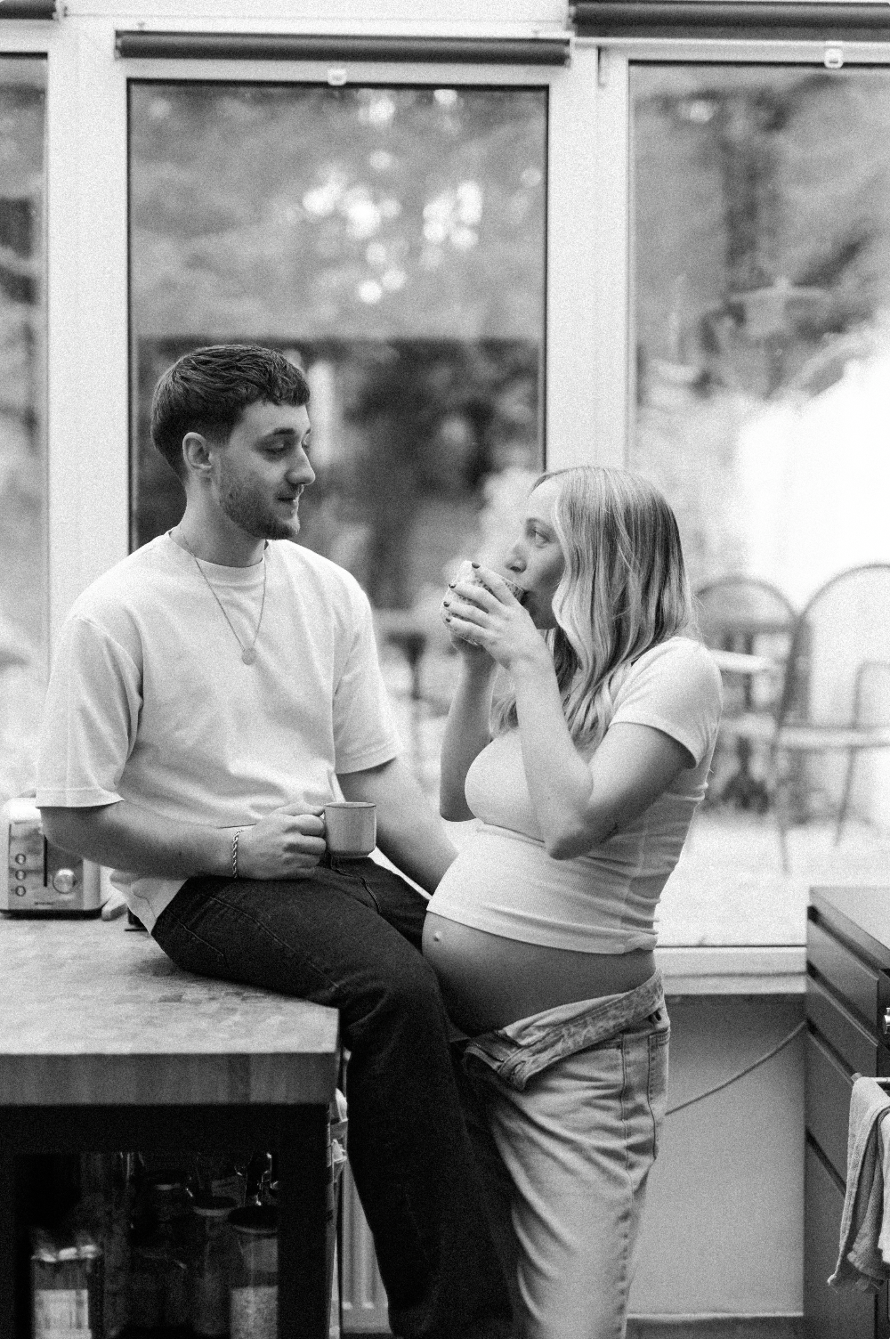 Deux jeunes adultes, un homme et une femme, dans une cuisine, partageant un moment complice avec des mugs, l'ambiance est chaleureuse et intime.