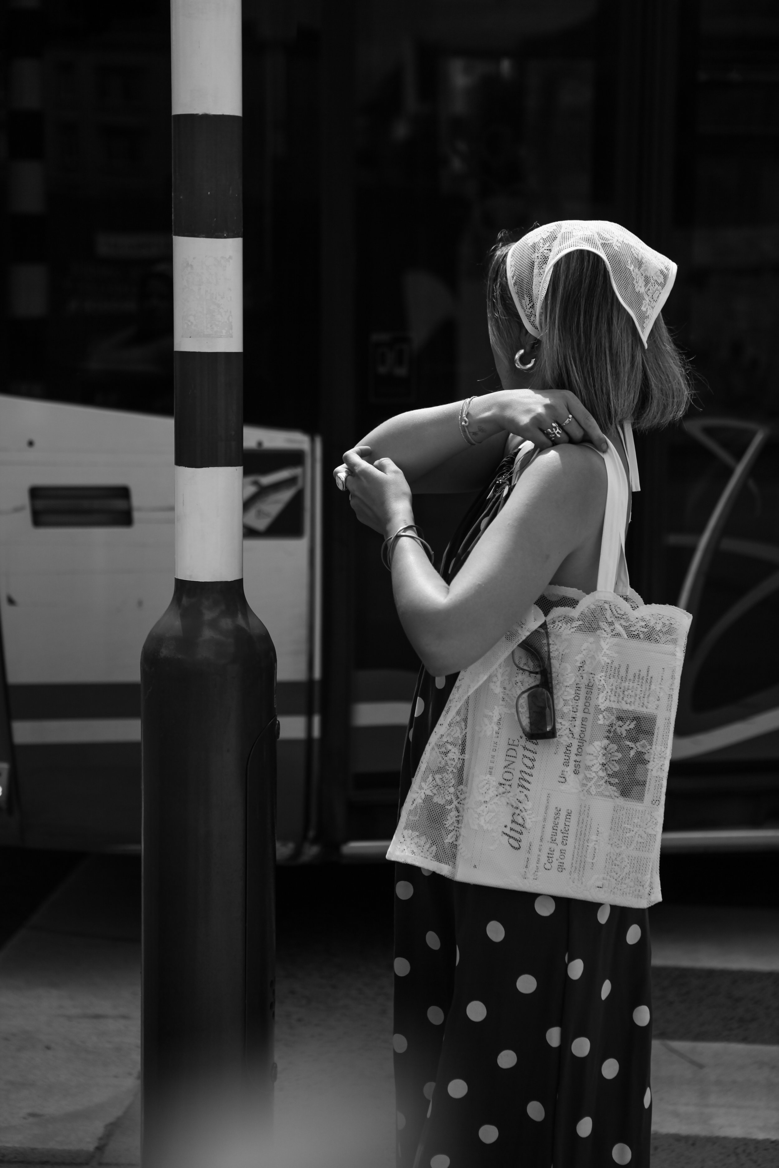 Une femme porte un sac en filet avec des motifs, appuyée contre une barrière de sécurité, portant une robe à pois, une casquette en dentelle, et des bijoux, en noir et blanc.