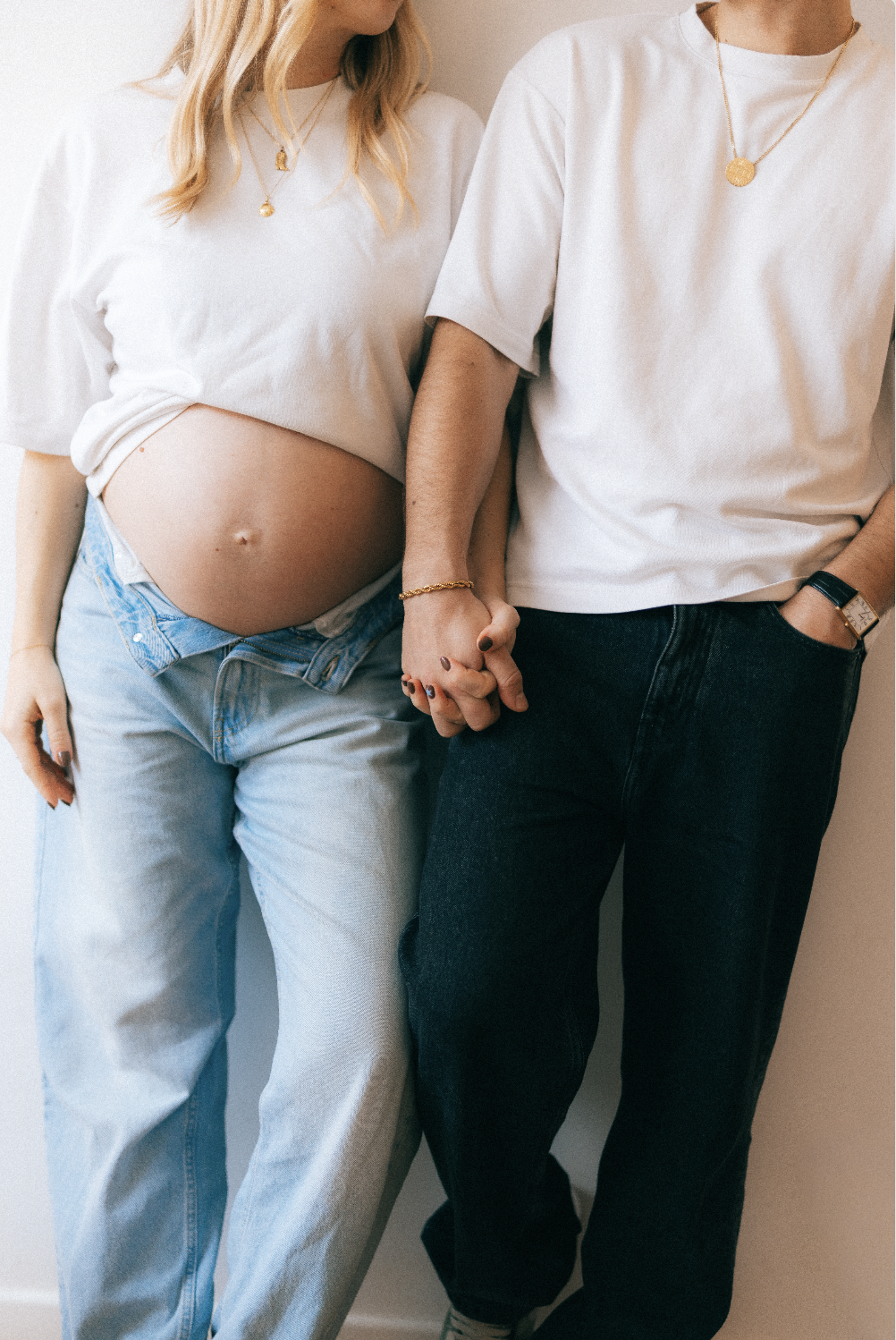 Une femme enceinte et un homme se tenant la main, tous deux portant des vêtements blancs et des jeans, leur arrestant au-dessus de la taille. La femme porte un t-shirt blanc noué au-dessus de son ventre arrondi et un pendentif en or. L'homme porte un