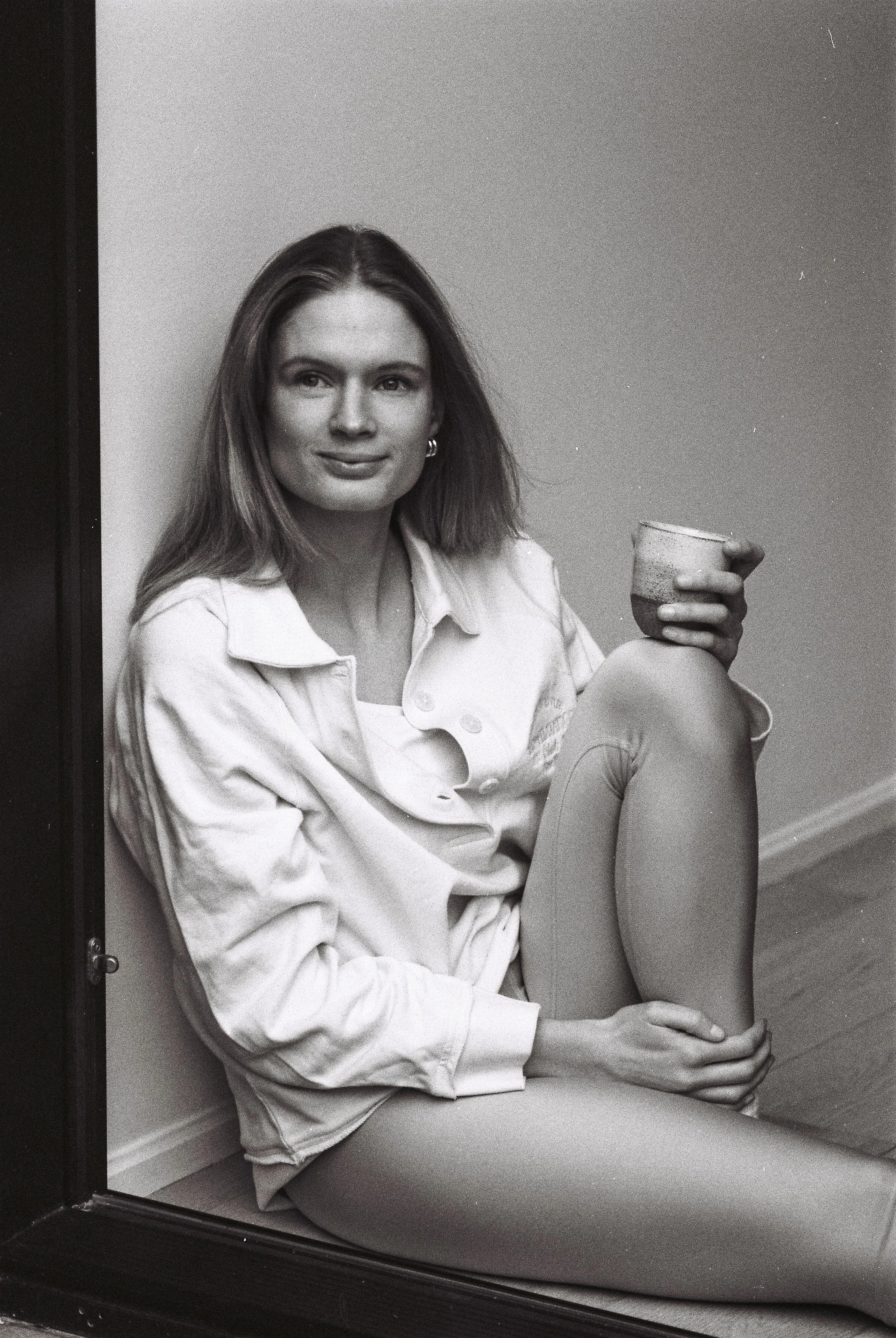 Une femme assise par terre dans un coin, tenant un bol, portant des collants et une veste claire, souriant légèrement, en noir et blanc.