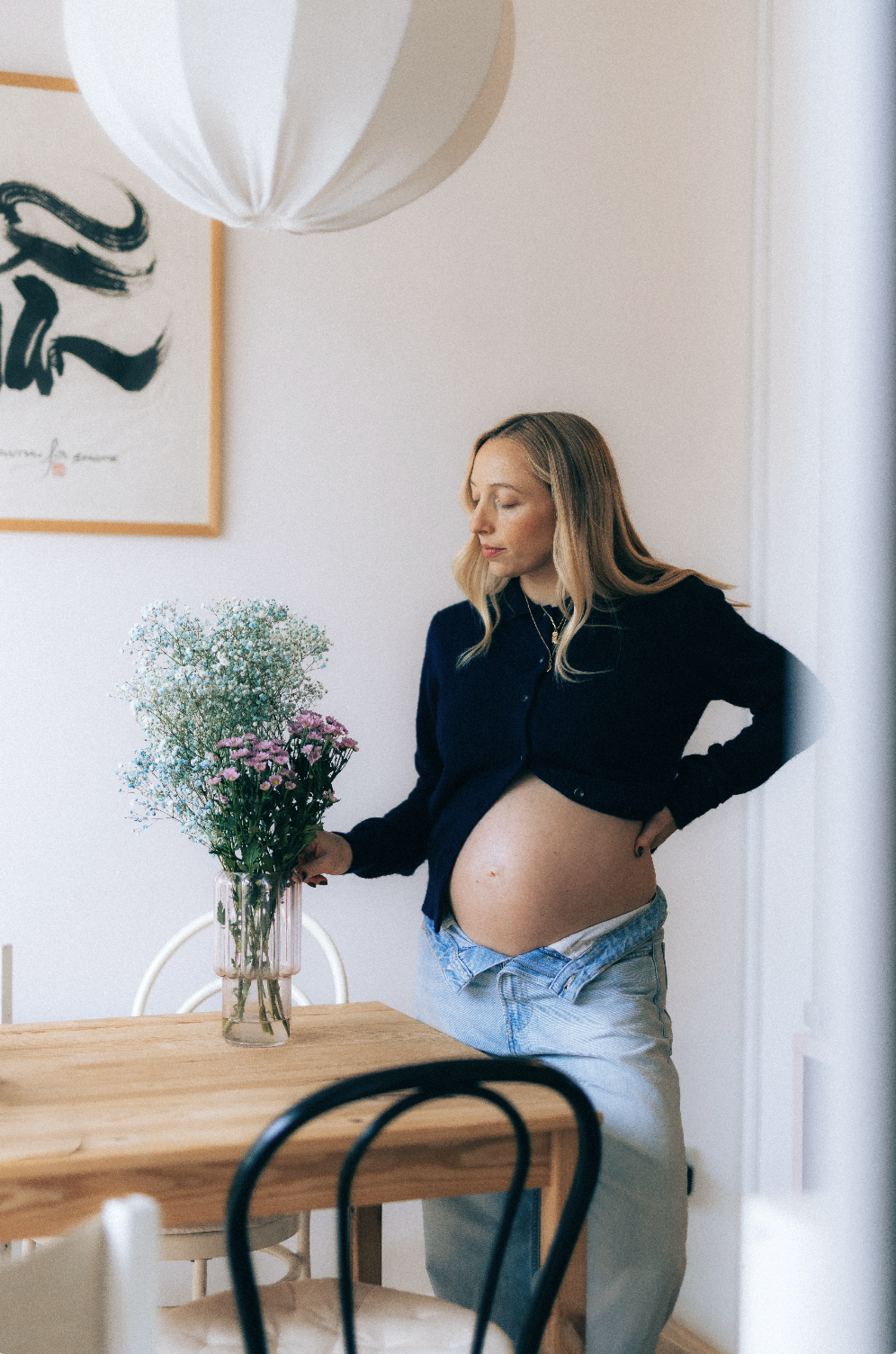Une femme enceinte regarde un bouquet de fleurs dans un vase en verre sur une table en bois dans une pièce lumineuse.