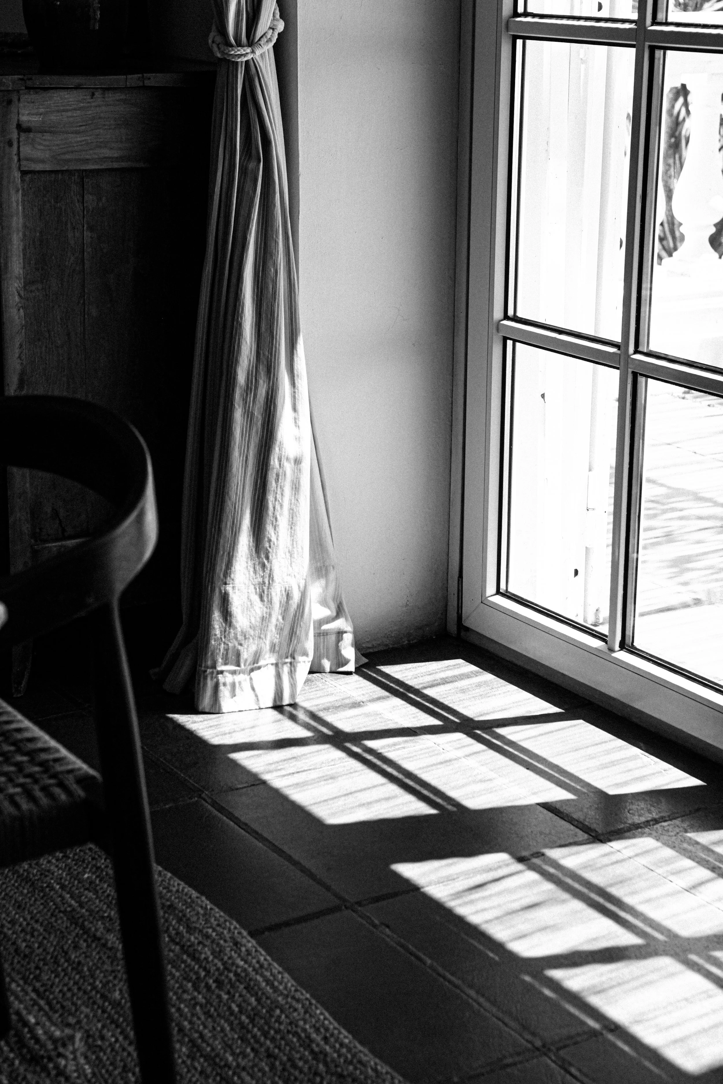 Lumière du soleil passant par une porte-fenêtre en verre, créant des ombres rectangulaires sur le sol en carreaux. Draperie à rayures sur le côté gauche.