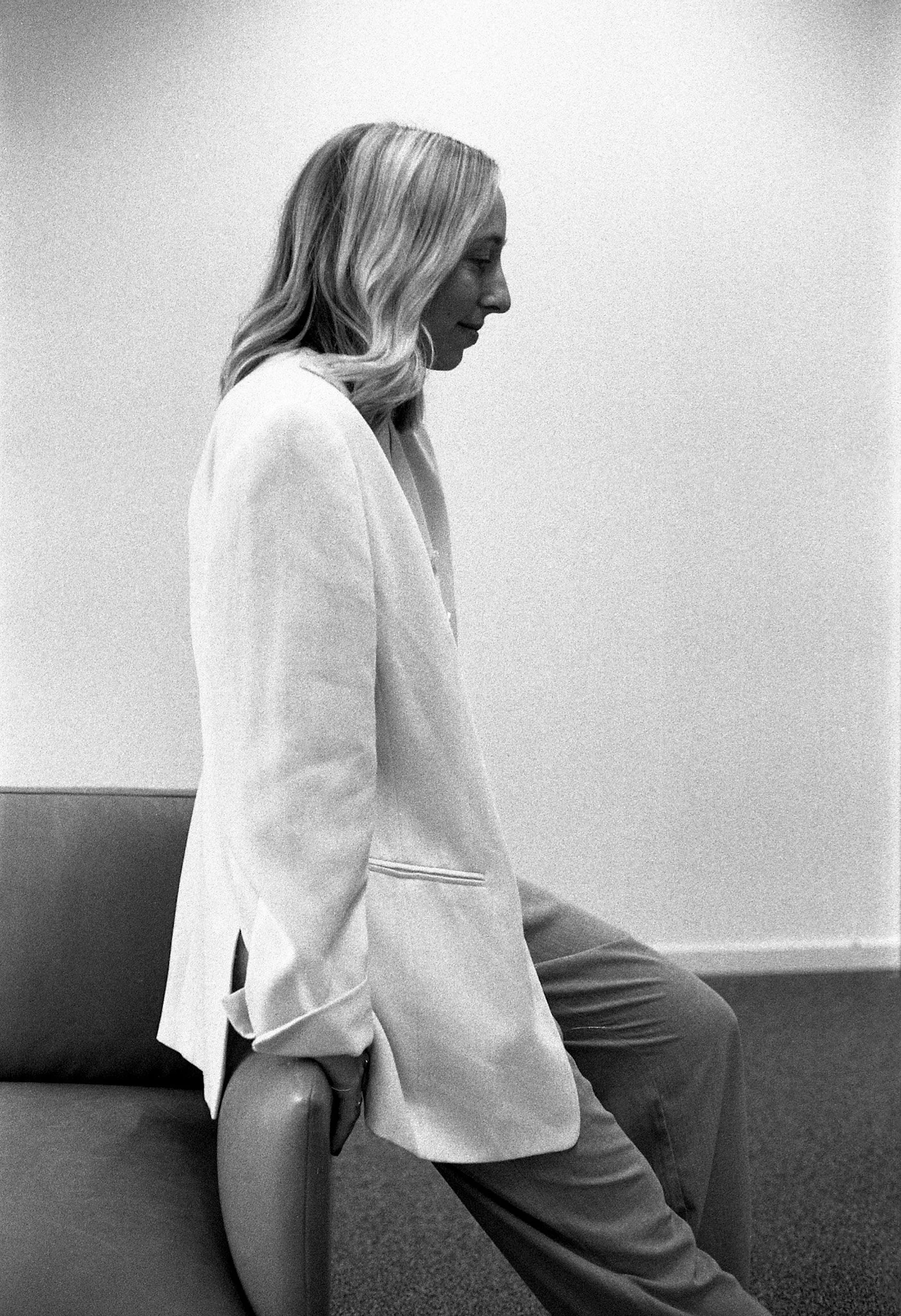 Une femme assise, vue de profil, portant un blazer clair et un pantalon, dans une pièce avec un mur blanc et un sol en moquette.