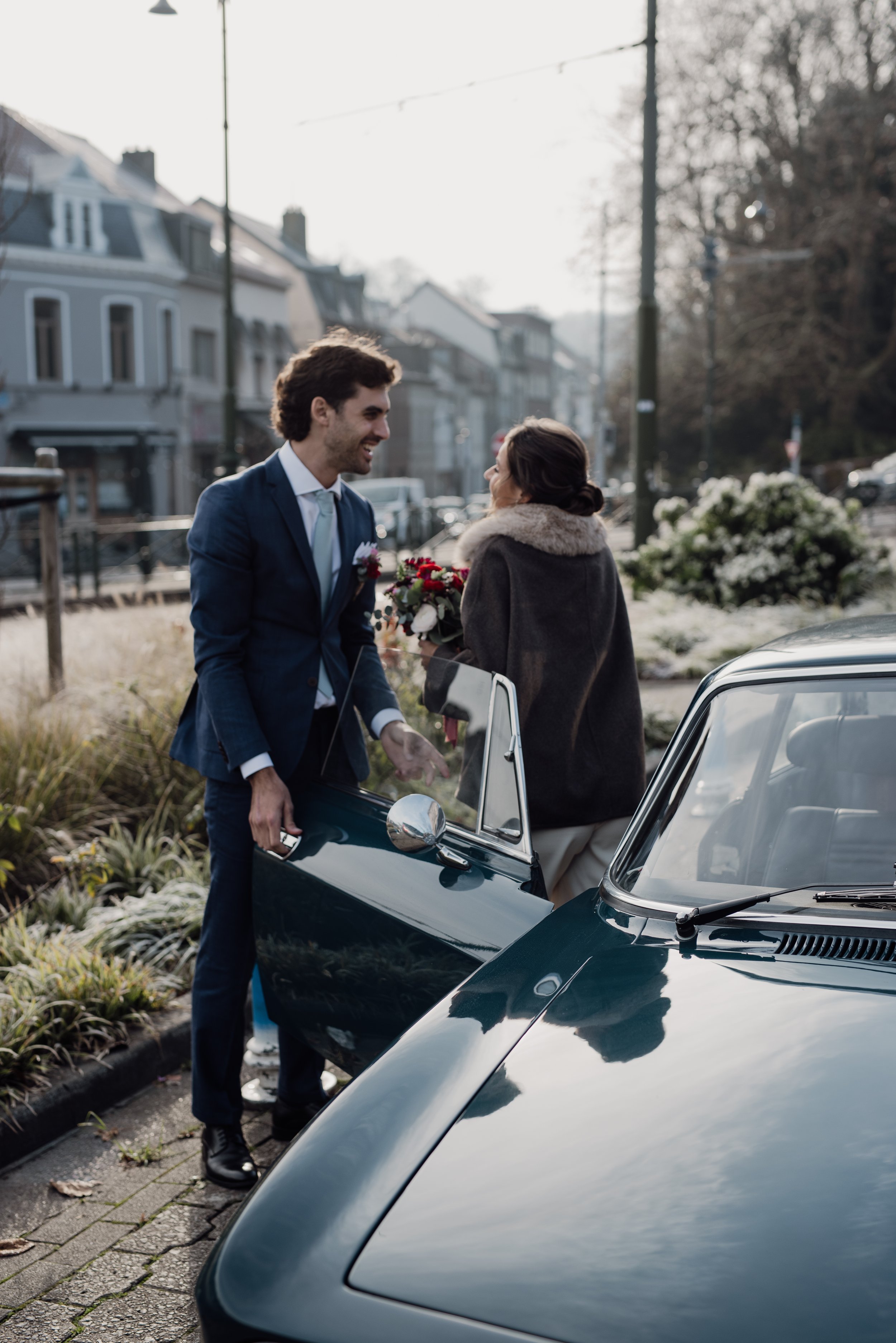 Un homme en costume de mariage souriant à une femme tenant un bouquet de fleurs, à côté d'une voiture noire dans une rue résidentielle.
