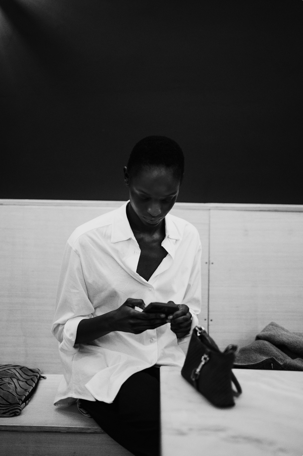 Une personne assise sur un banc, regardant son téléphone, portant une chemise blanche avec des vêtements à côté.