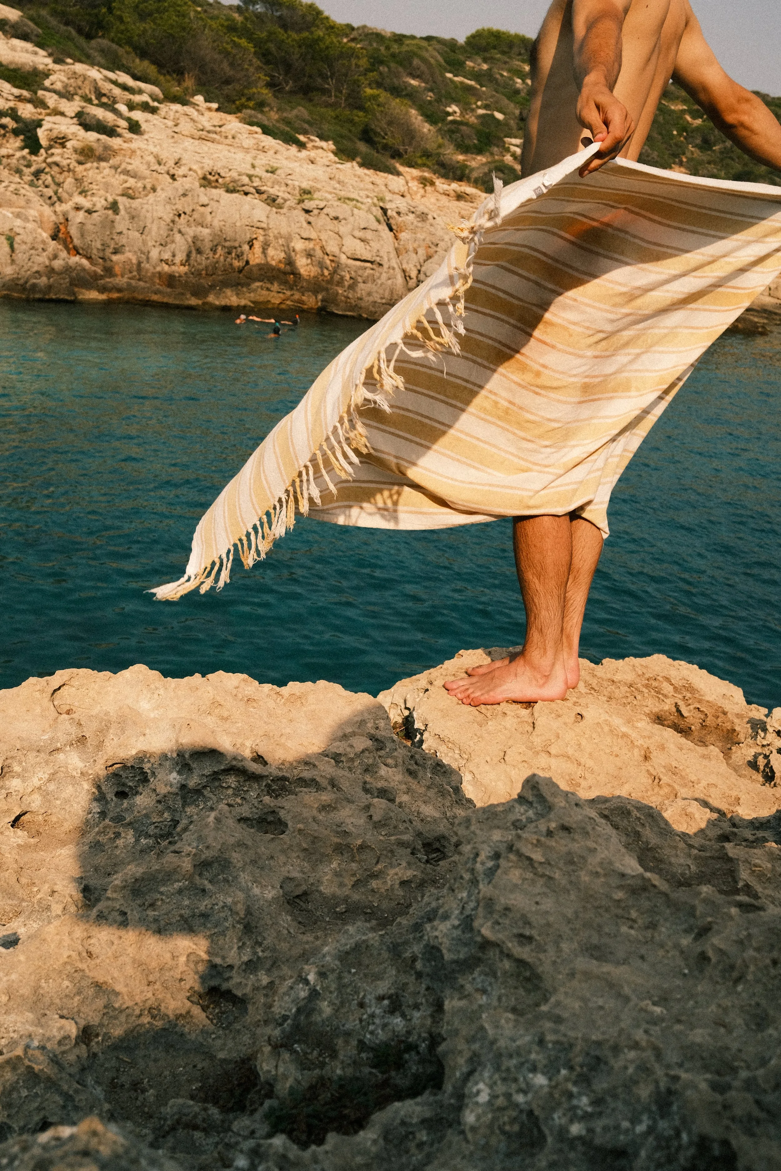 Une personne debout sur une roche près de la mer, tenant une serviette ou un tissu léger.
