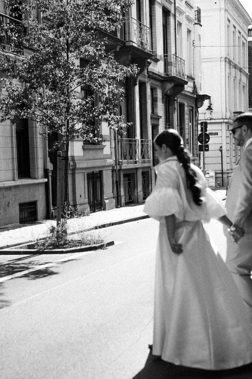 Un couple en vêtements de mariage marche main dans la main dans une rue urbaine. L'image est en noir et blanc. Il y a des bâtiments résidentiels avec balcons et une lampe à droite.